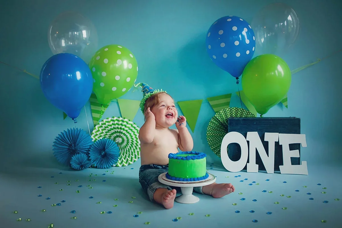 One-year-old baby celebrating a milestone birthday with a blue and green cake smash setup and balloons in a photography studio