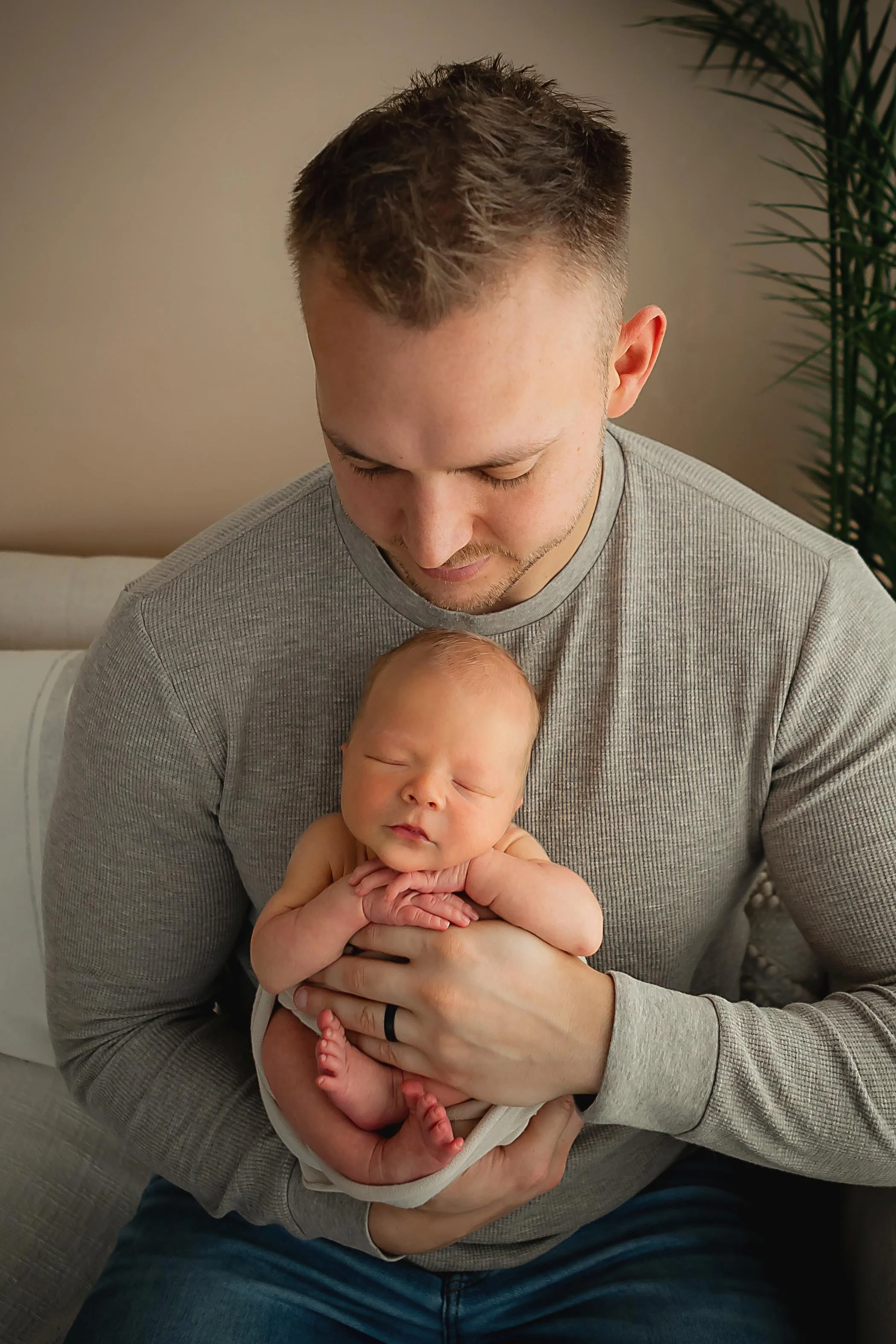 Dad in a gray shirt holding his baby boy facing outward with baby's hands resting peacefully on Daddy's hand showing Daddy's wedding band