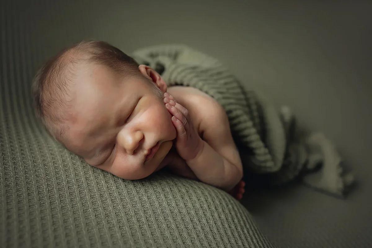 Newborn baby boy sleeping on his side with his hand resting on his cheek with a sage green wrap.