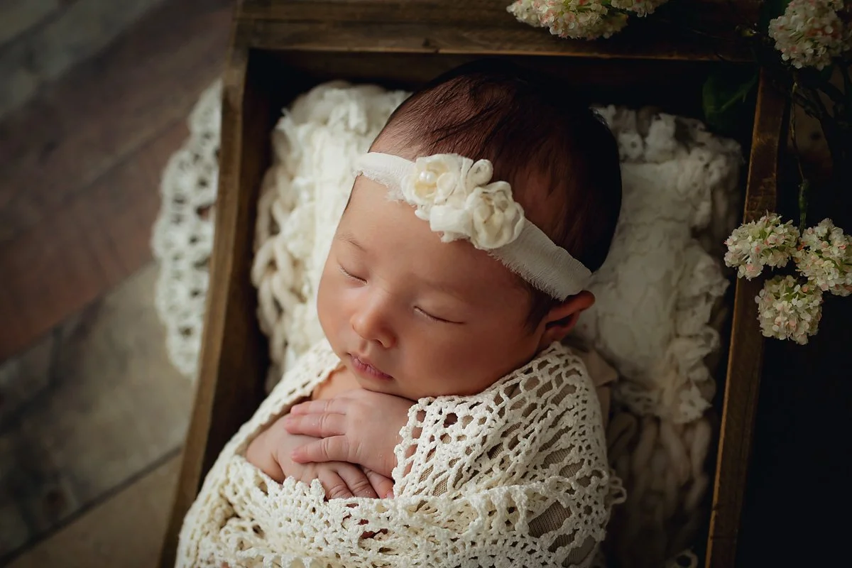 Close-up of a sleeping newborn girl wrapped in cream crochet lace with a delicate rosette headband, nestled in a wooden crate by Memory Lane Photography Indianapolis.