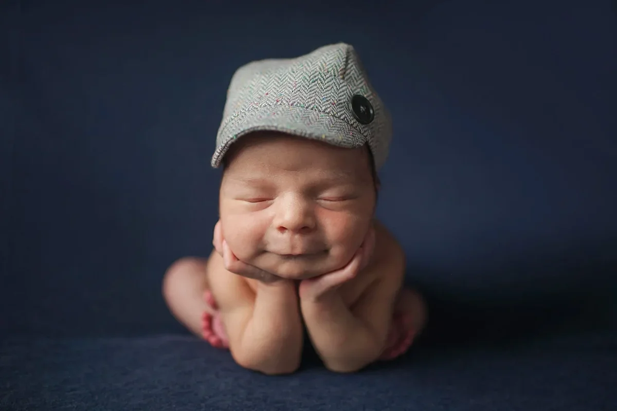 Newborn baby boy posed froggy style with his head resting in his hands smiling with a cute little hat on.