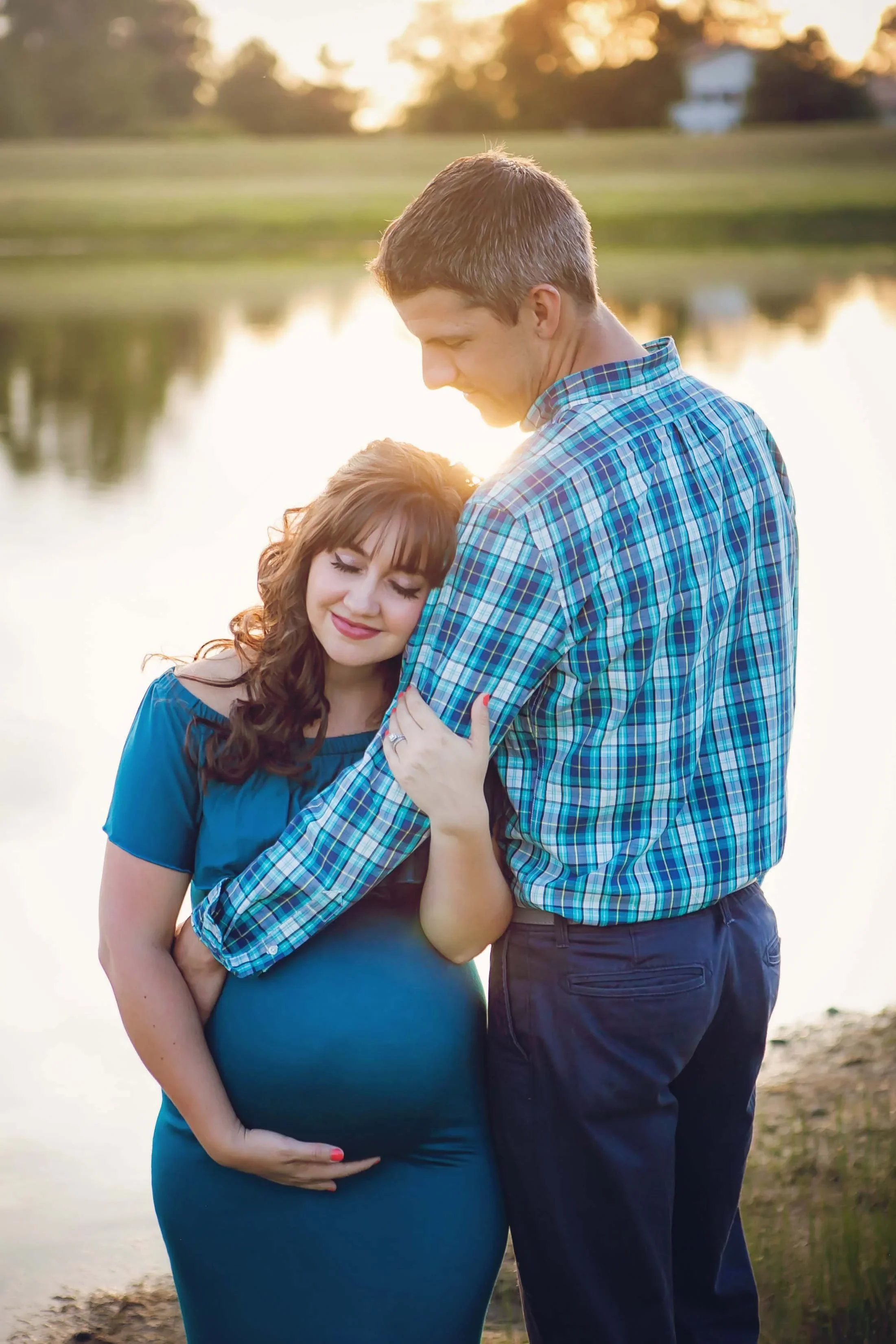 A pregnant woman and a man embracing outdoors near a lake at sunset, with the woman smiling and resting her head on the man's chest.