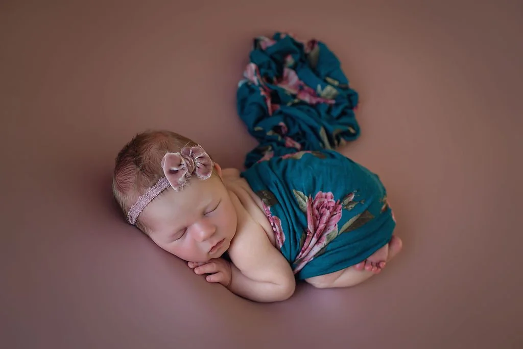 Mauve background with baby girl sleeping on her tummy wearing a mauve velvet bow and covered with a floral wrap for her newborn photography session.