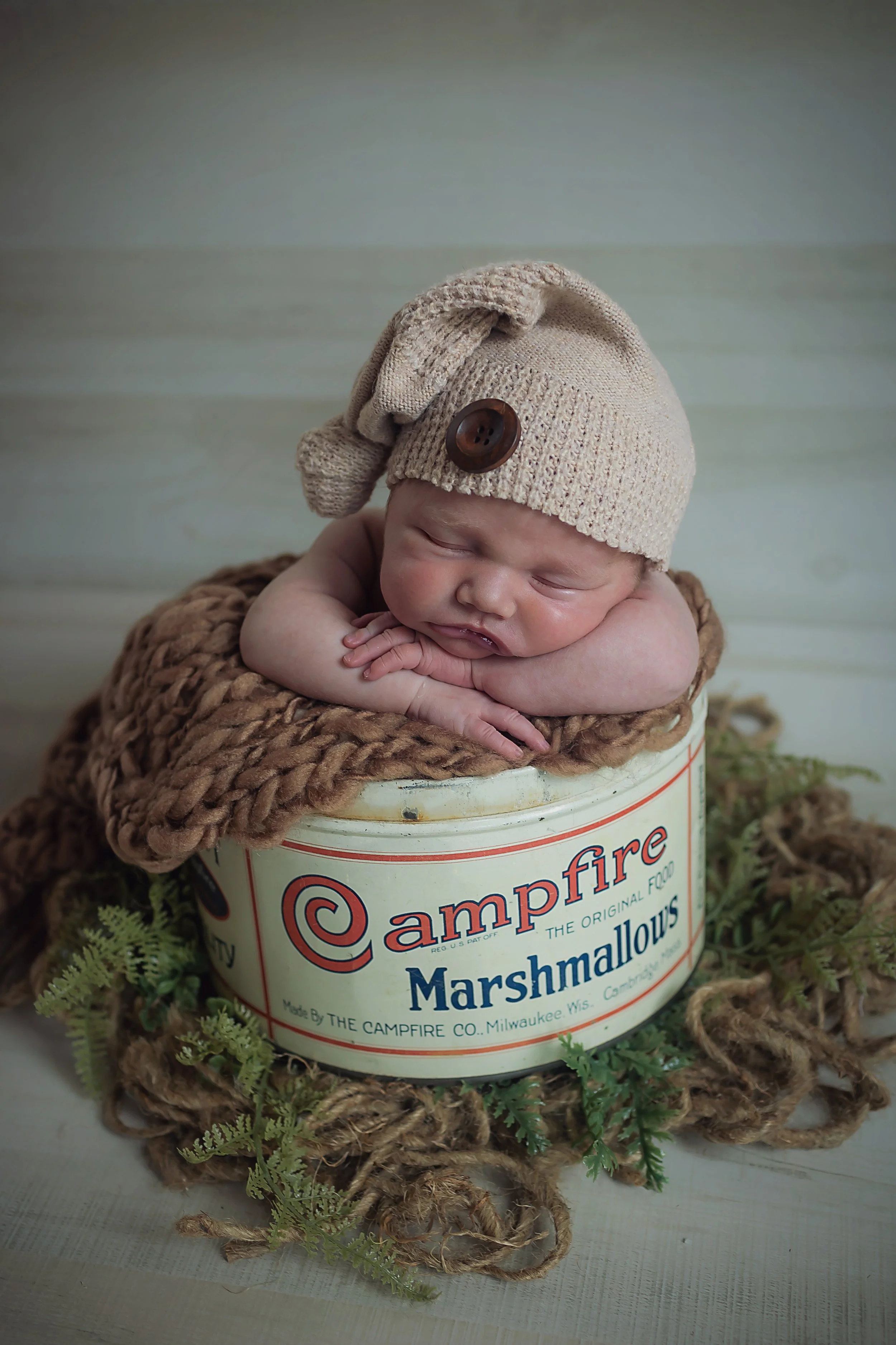 Newborn wearing a beige sleepy hat snuggled in a vintage Campfire Marshmallows tin sleeping propped up on their arms.