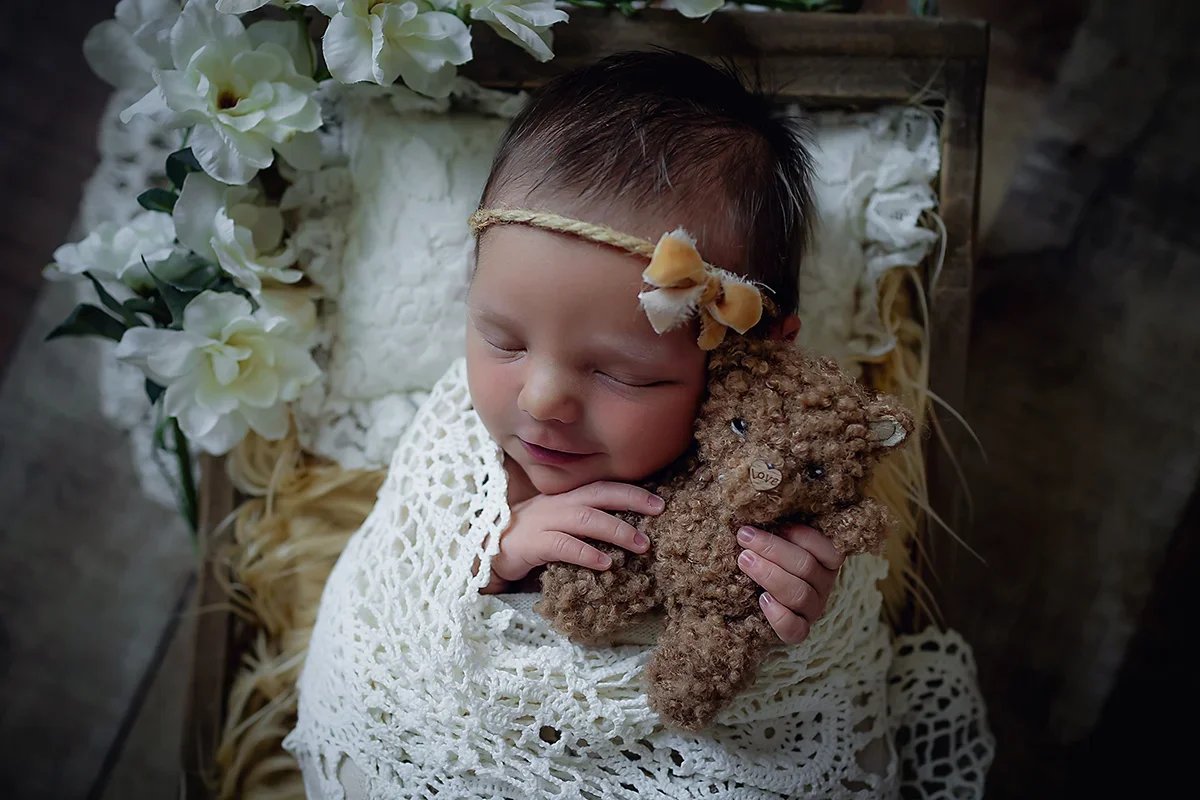 Smiling newborn baby wrapped in a lace wrap holding a little teddy bear with a heart button nose.