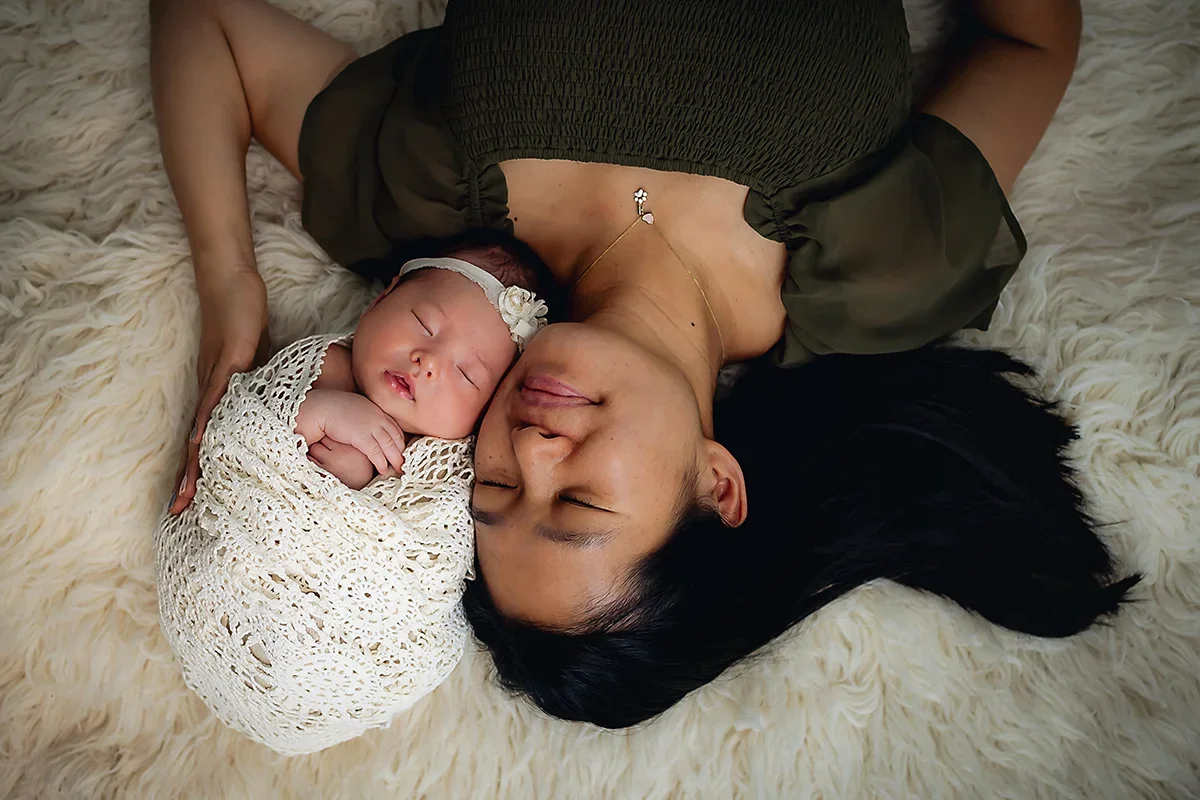 Newborn baby laying with mom next to her  on a white fluffy rug with baby in a lace wrap.