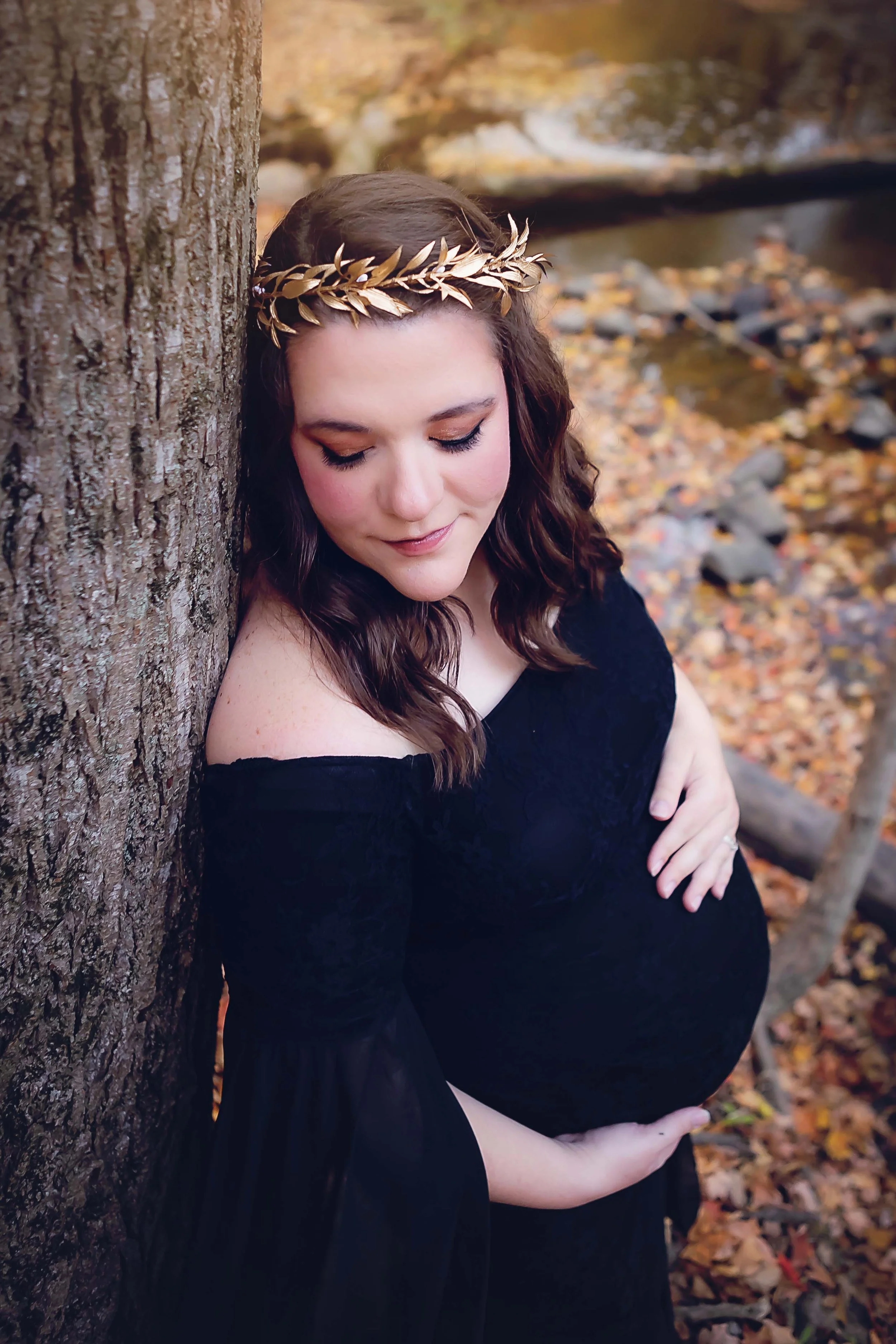 Expecting mother posing for her maternity portrait  in a black gown with a golden leaf crown with fall foliage as her backdrop at a creek in an  Indianapolis park
