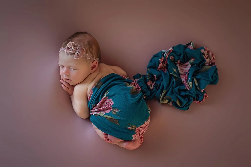 Newborn baby girl on mauve background with floral wrap covering her, wearing a mauve velvet bow during her newborn photography session in Indianapolis.