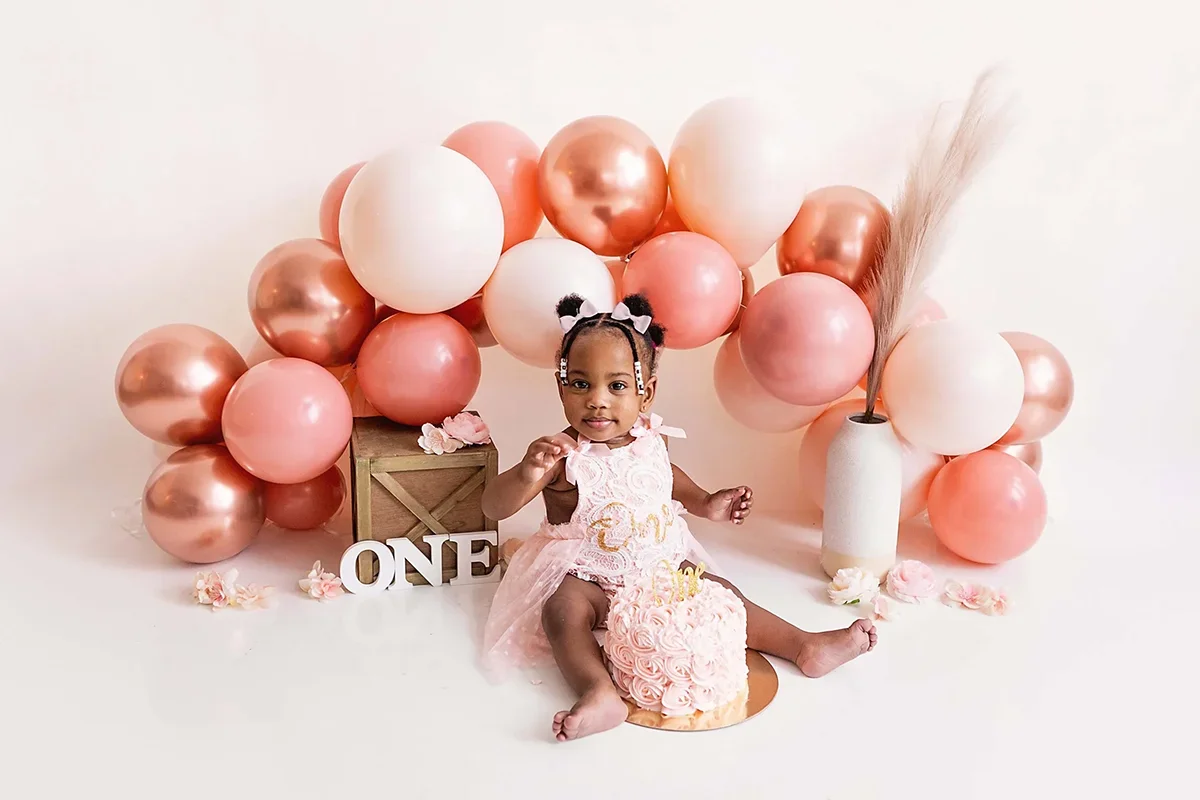 One year old birthday photoshoot, little girl in pink lace dress sitting behind a smash cake with a pink balloon arch and the letters ONE behind her.