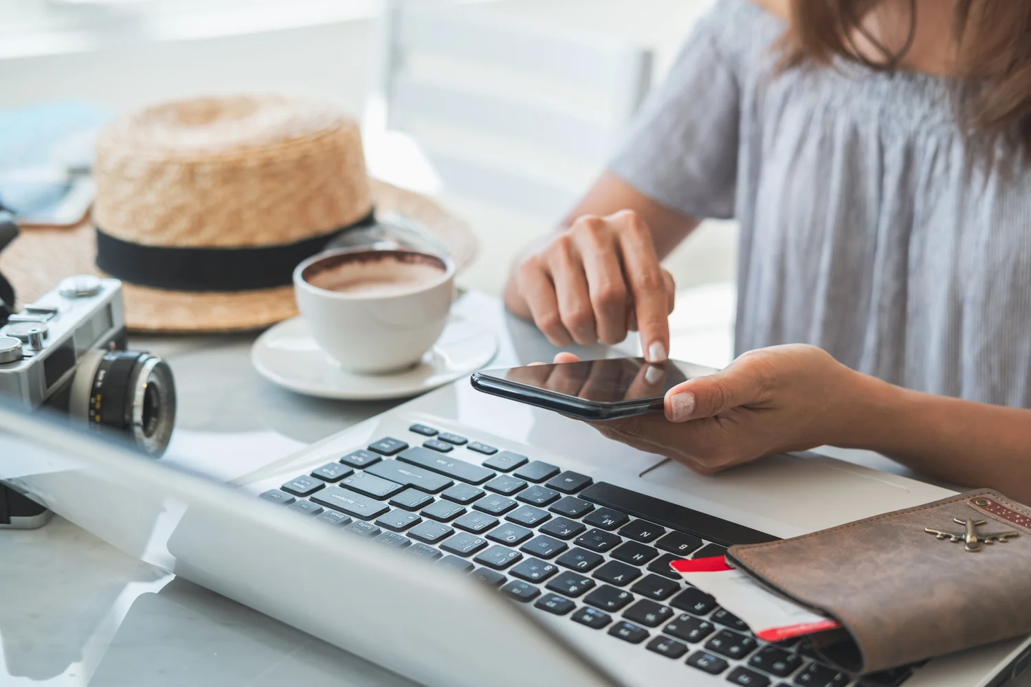 Young woman searching for flights and accommodation on her smartphone and laptop. A cup of coffee, hat, camera and passport are also on the table.