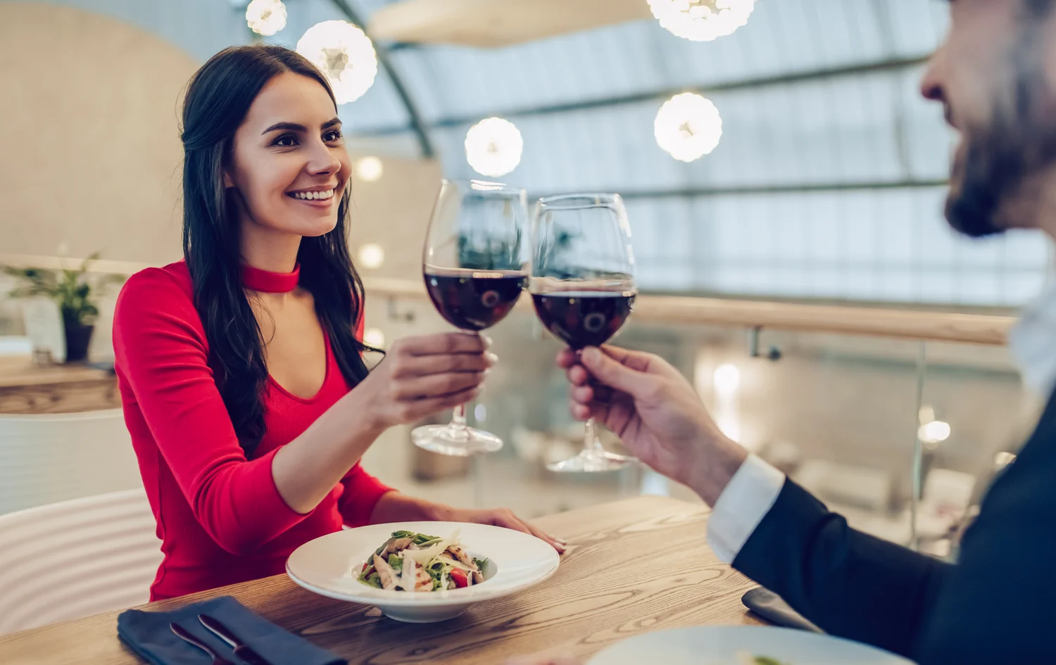 A young brunette woman in red and her male date clinking glasses of red wine at a fine dining restaurant