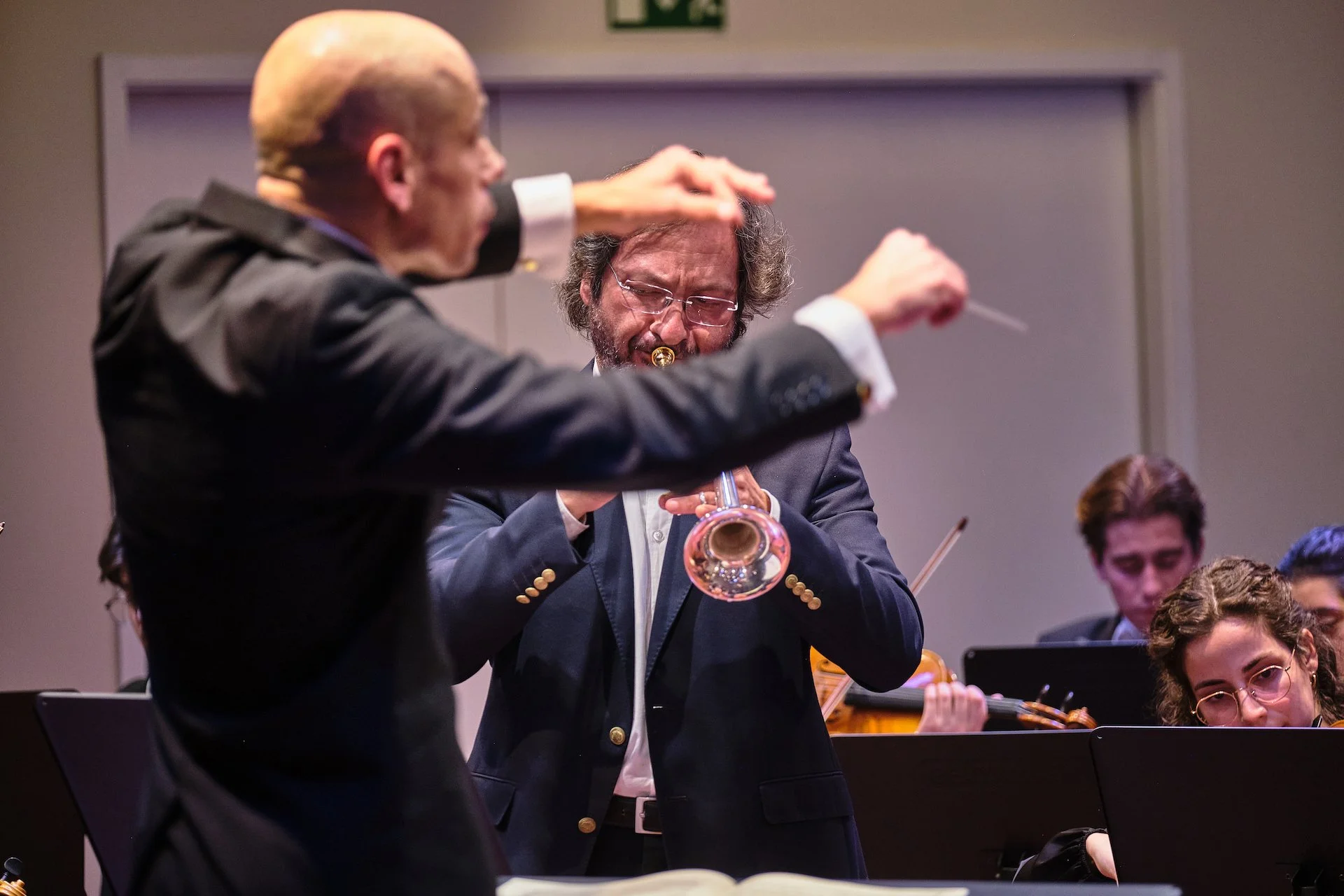 Orchestra conducting and playing with a conductor in the foreground and a woman and other musicians in the background.