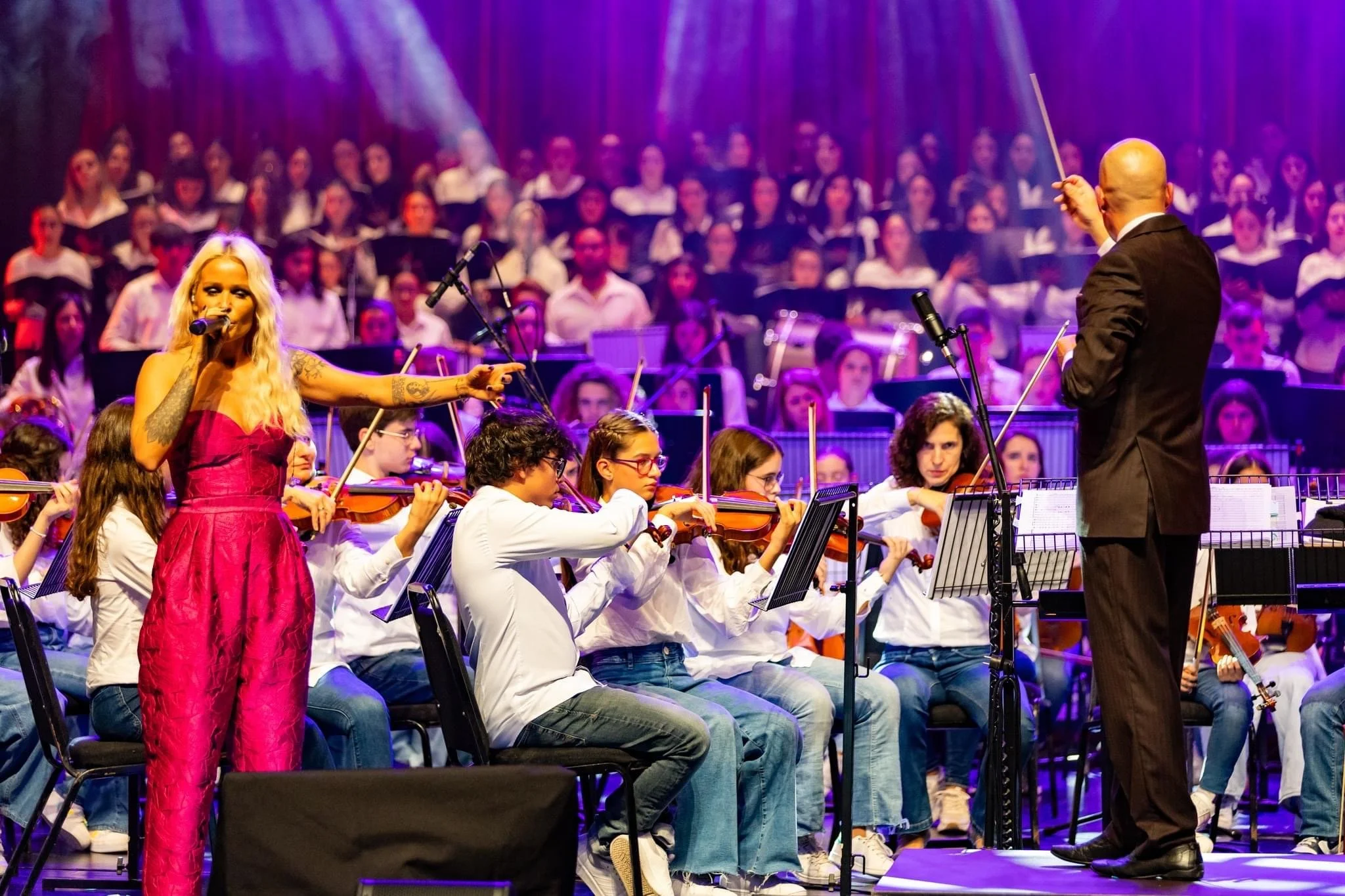 A live orchestral performance with a singer in a pink dress and a conductor leading the orchestra on stage with a large choir in the background.