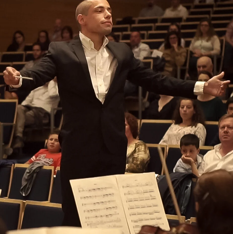 A man in a black tuxedo and white shirt conducting an orchestra in front of an audience.