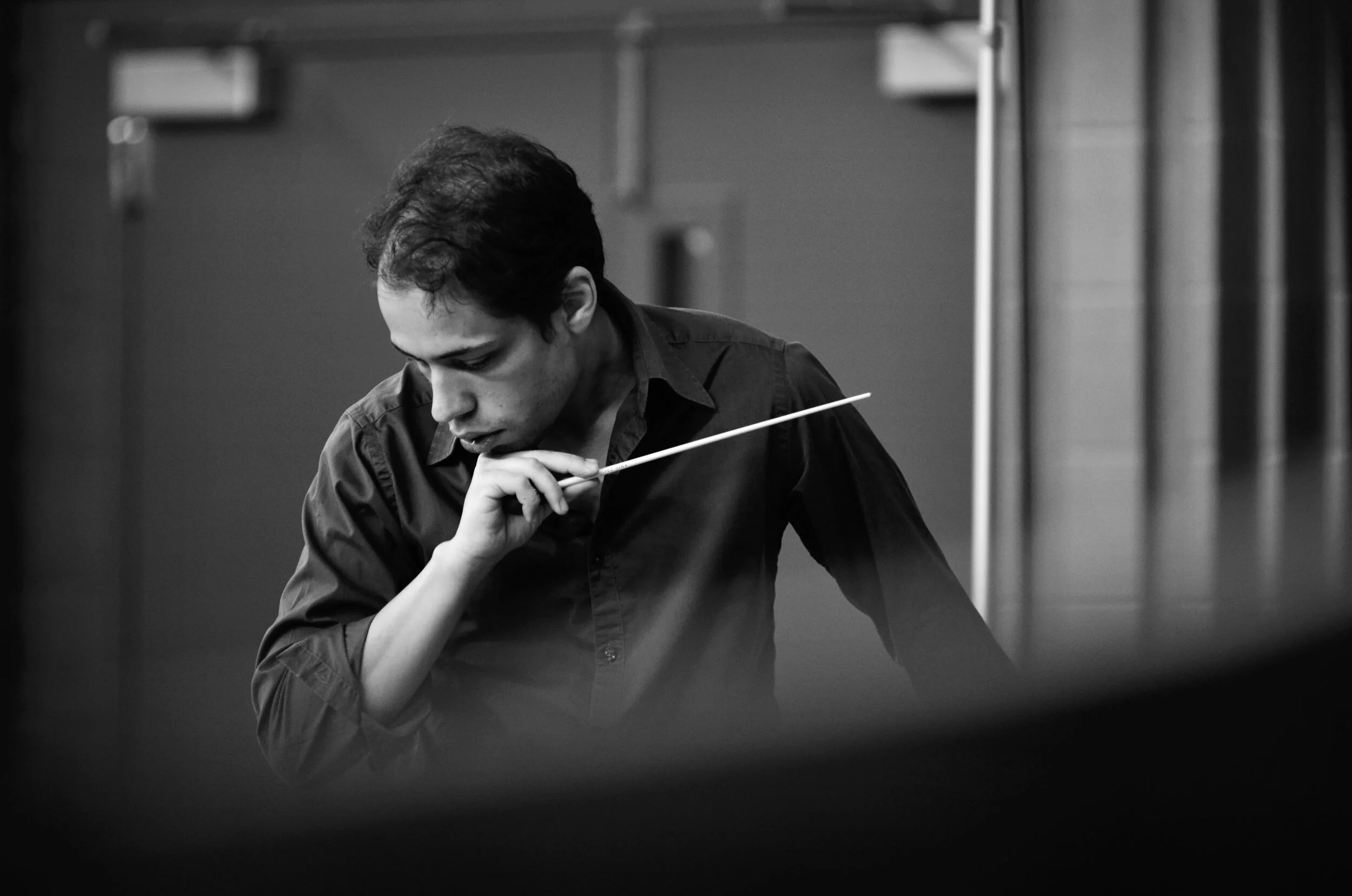 Black and white photograph of a man holding a conductor's baton, carefully examining it in a music rehearsal or performance setting.