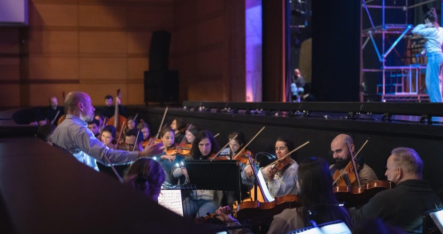Orchestra rehearsing in a concert hall, with the conductor leading and musicians playing violins.