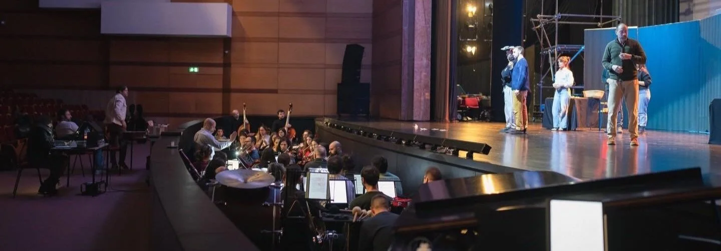 People on stage performing or rehearsing, with a crew member overseeing from the floor. Down in the audience or control area, people working with computers and equipment, preparing or managing the performance. The setting appears to be a theater or a