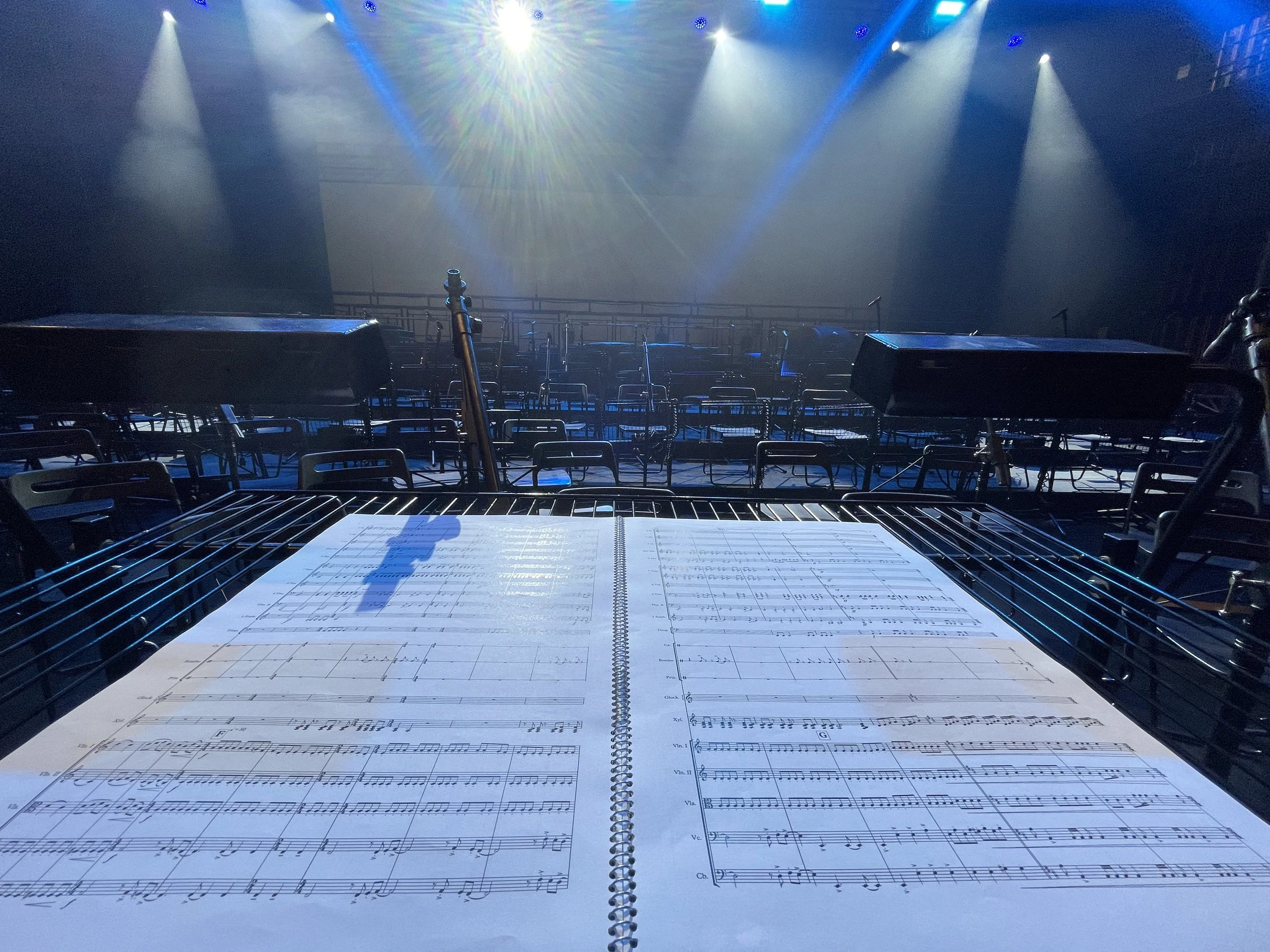 An empty stage with rows of chairs, microphones, and a large screen in the background, taken from behind a music stand with sheet music, in a well-lit concert hall or theater.