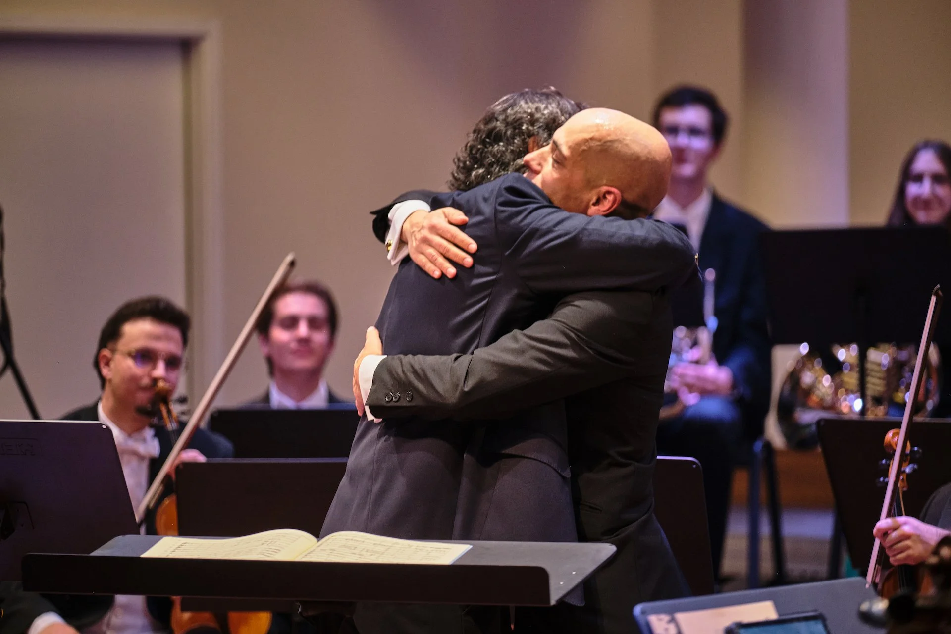 Two men hugging warmly in front of an orchestra, with several musicians observing in the background.