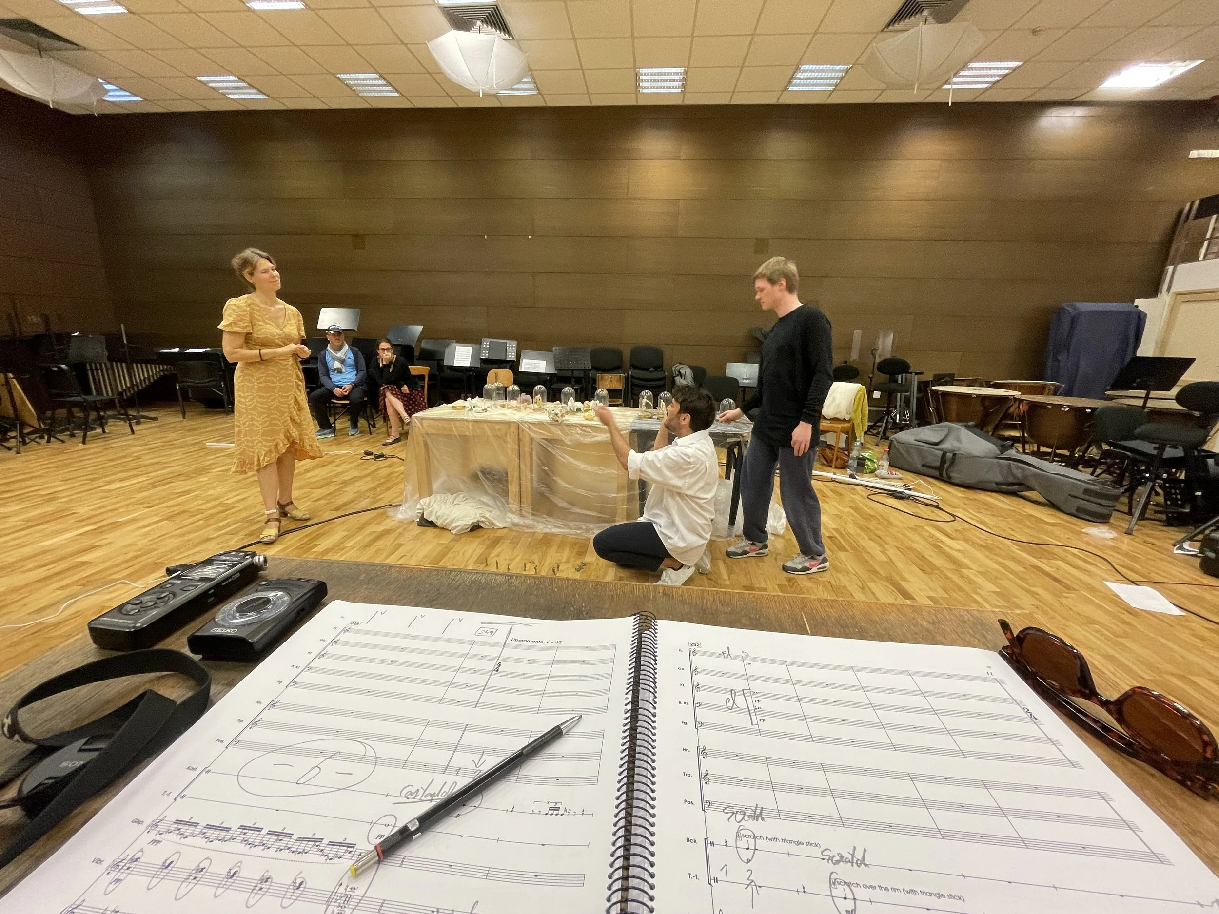 A spacious music rehearsal room with a wooden floor and wall. Several people are sitting and standing near a table with transparent plastic covering, surrounded by musical instruments and chairs. In the foreground, there is sheet music, a pen, sungla