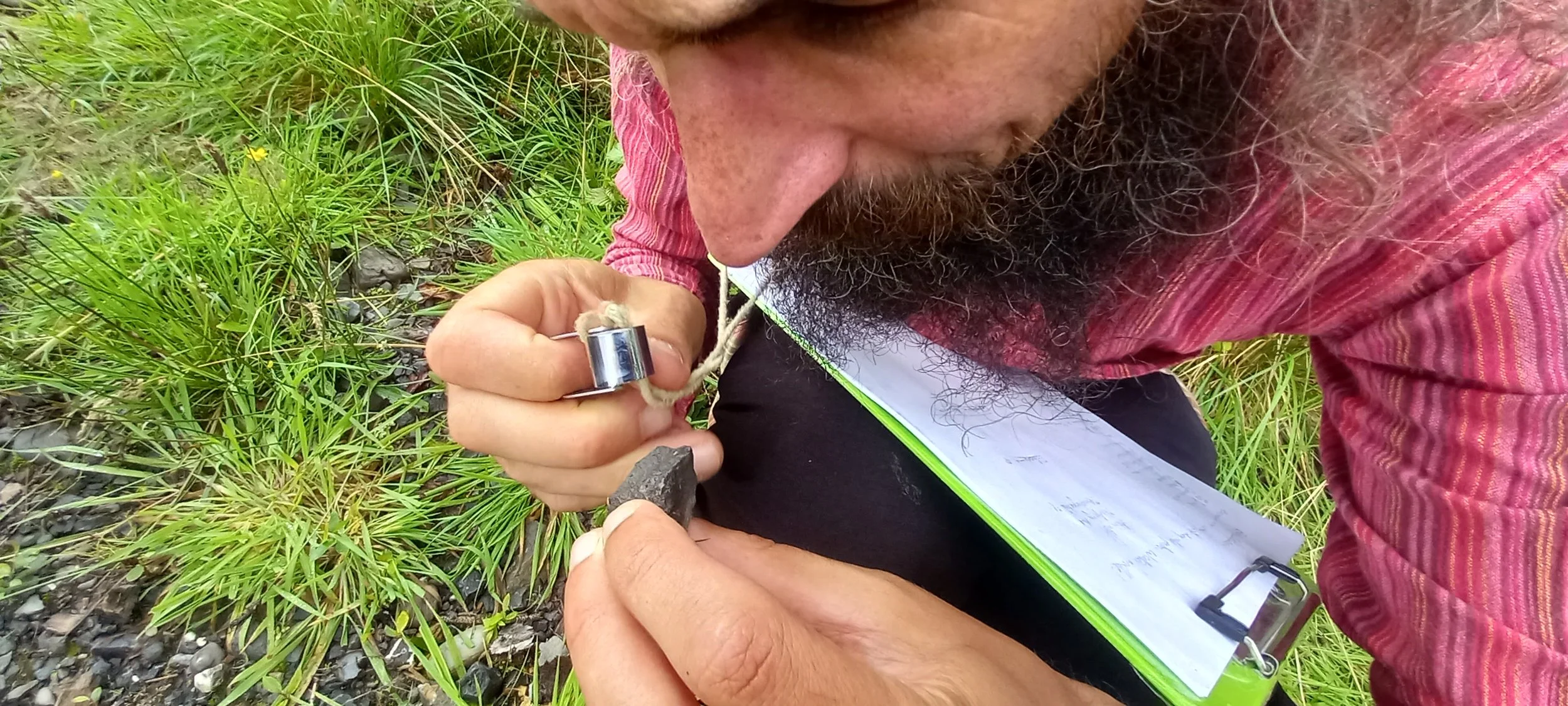 A close up of a mans face and his bear. He has a tiny magnifyer in his hand and is looking at a tiny frog in the woodland.