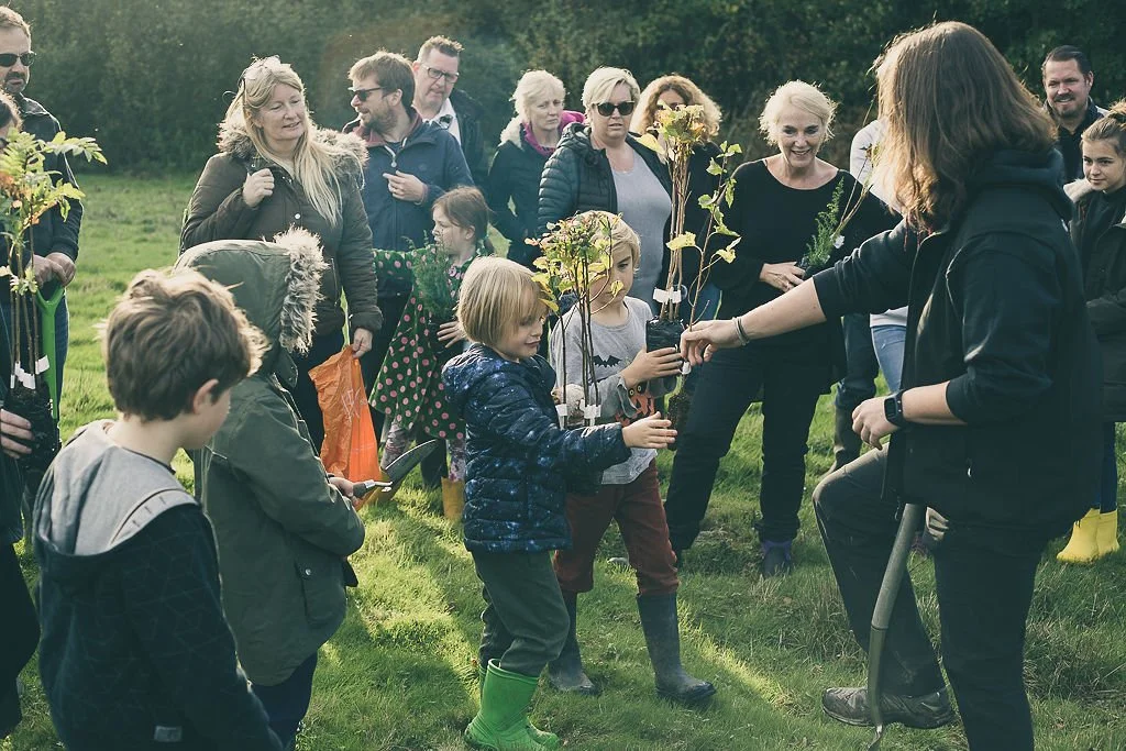 A group of people standing in a semi cricle in a field with trees lining the fild in the background. Young children are in the foreground hold saplings and an adult is talking to them about the trees.