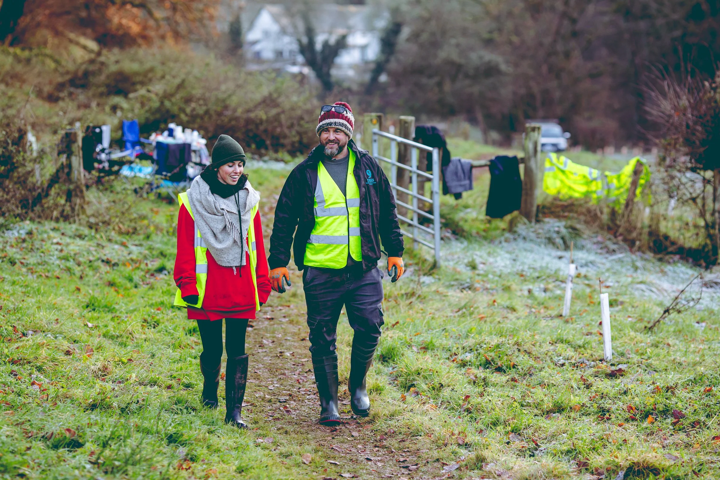 A woman in high vis, red jacket, wellies and beanie walking next to a man in high vis and a beanie. They are in the field at a tree planting site and smiling to camera
