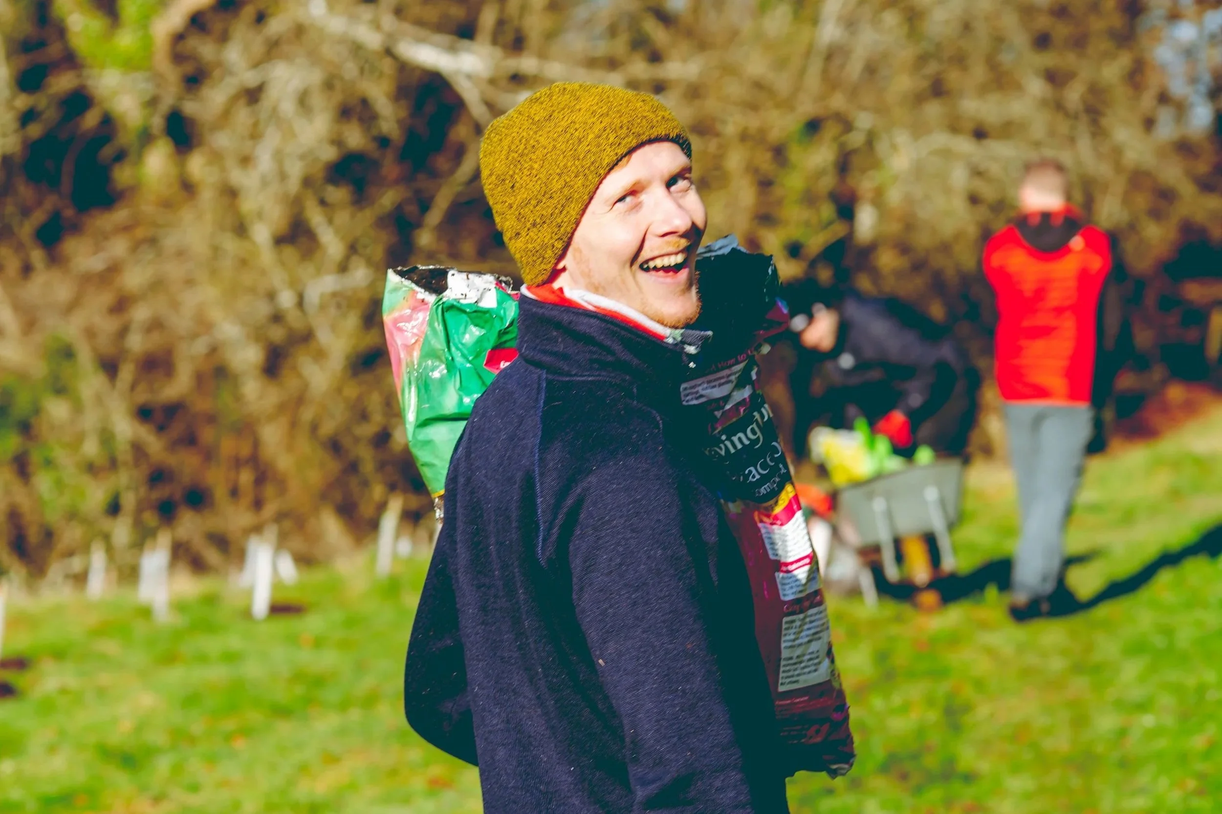 A Man standing in  field carrying a bag of mulch and he is half turning back around smling to camera