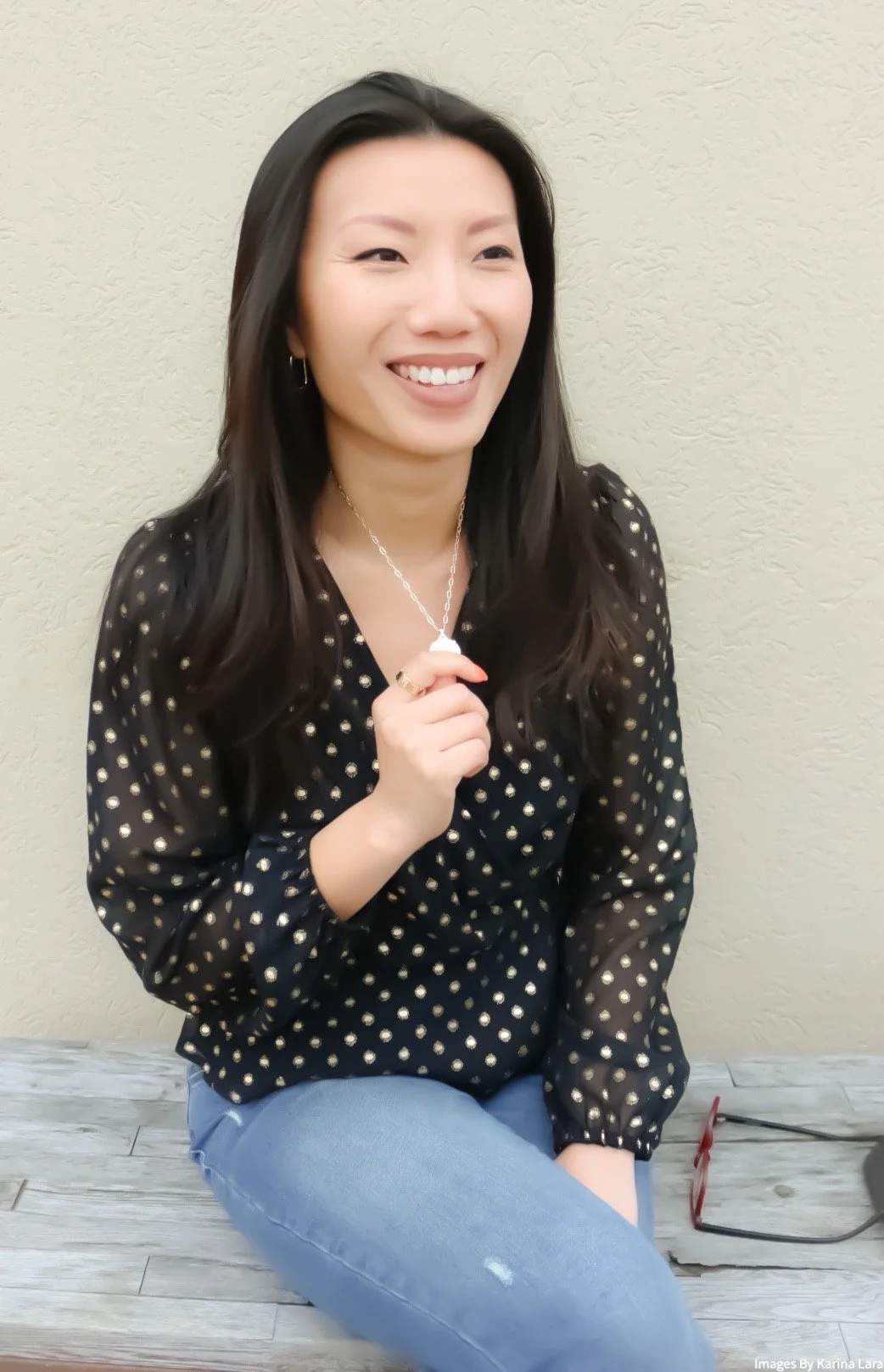 Anna sitting on a grey wooden bench, holding the pendant of her necklace between her index finger and thumb. She is smiling pleasantly.