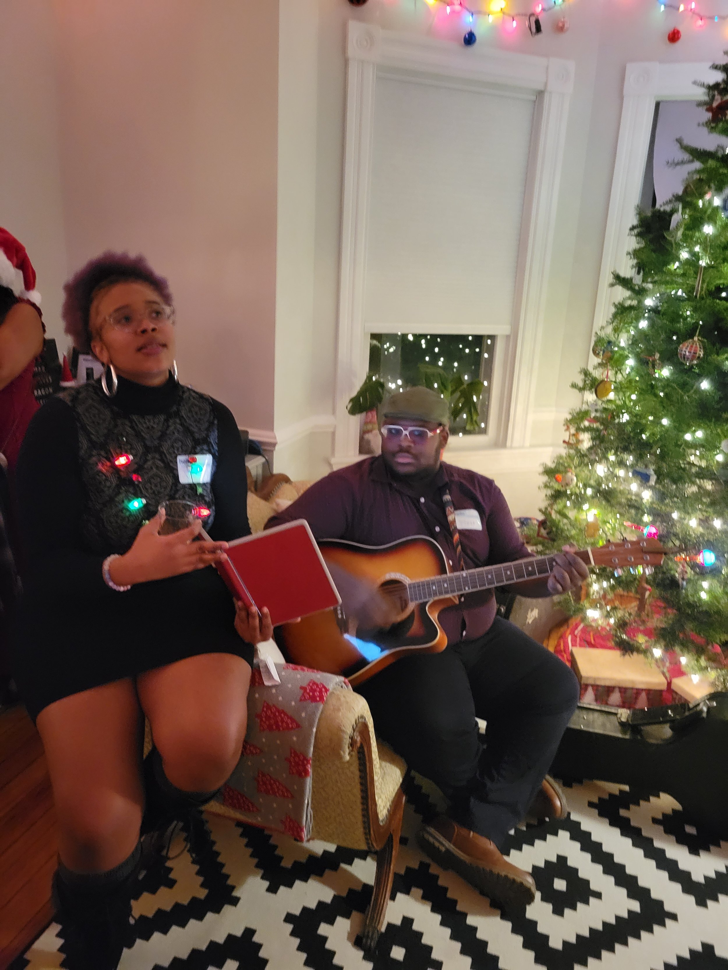    Vocal percussionist and Alto, Amber Leigh, holding up an iPad for our Executive Director, Vince, to read music from as he plays guitar for an instrument accompanied number at a private Christmas party Noteworthy performed at in 2022.   