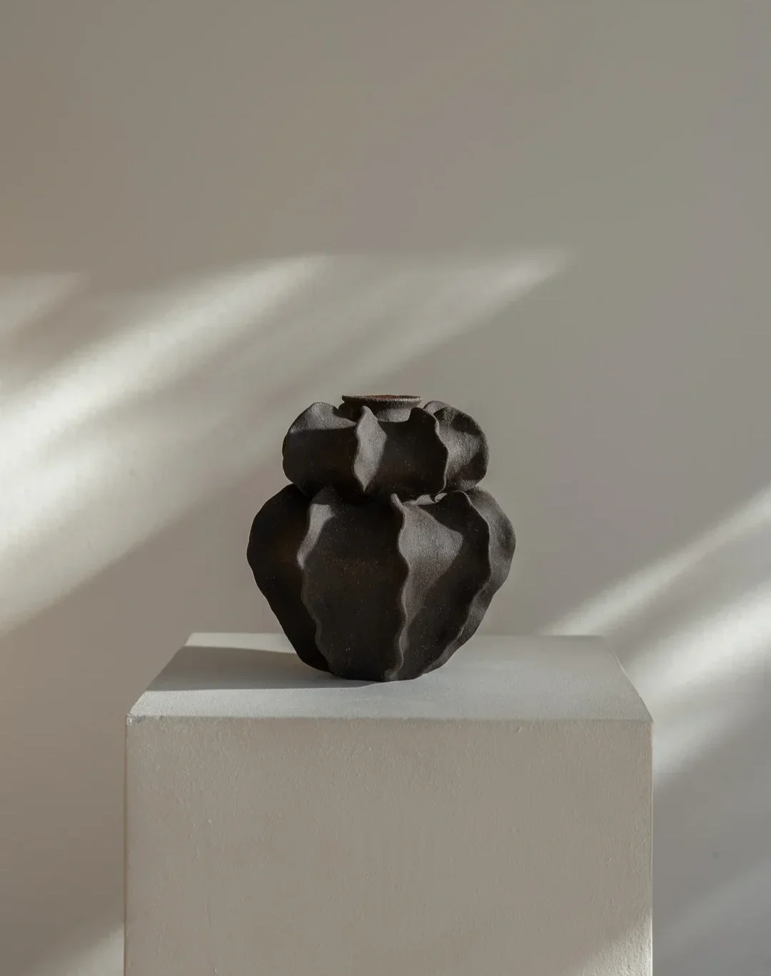 Ripple Vase — a handbuilt, sculptural ceramic vessel displayed on a pedestal, its dark textured form catching soft directional light.