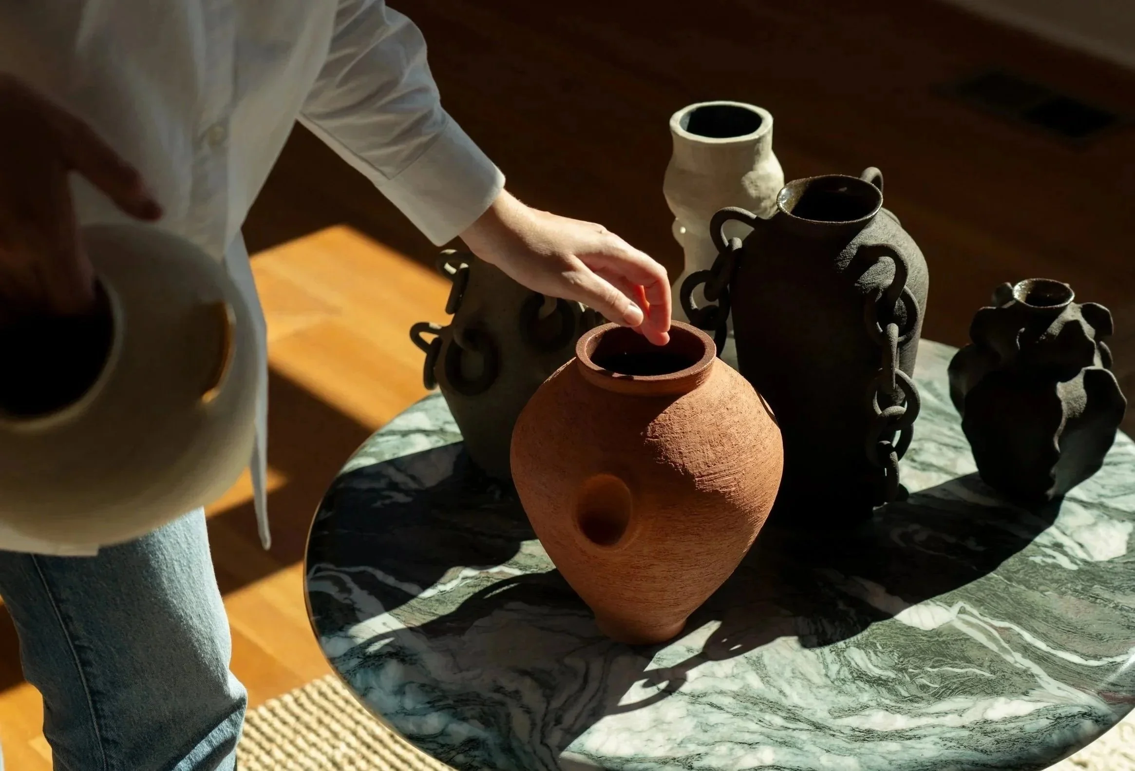An artist arranging a group of handbuilt Ruby Bell Studio sculptural ceramic vessels on a marbled coffee table, with sunlight highlighting the varied textures and forms.