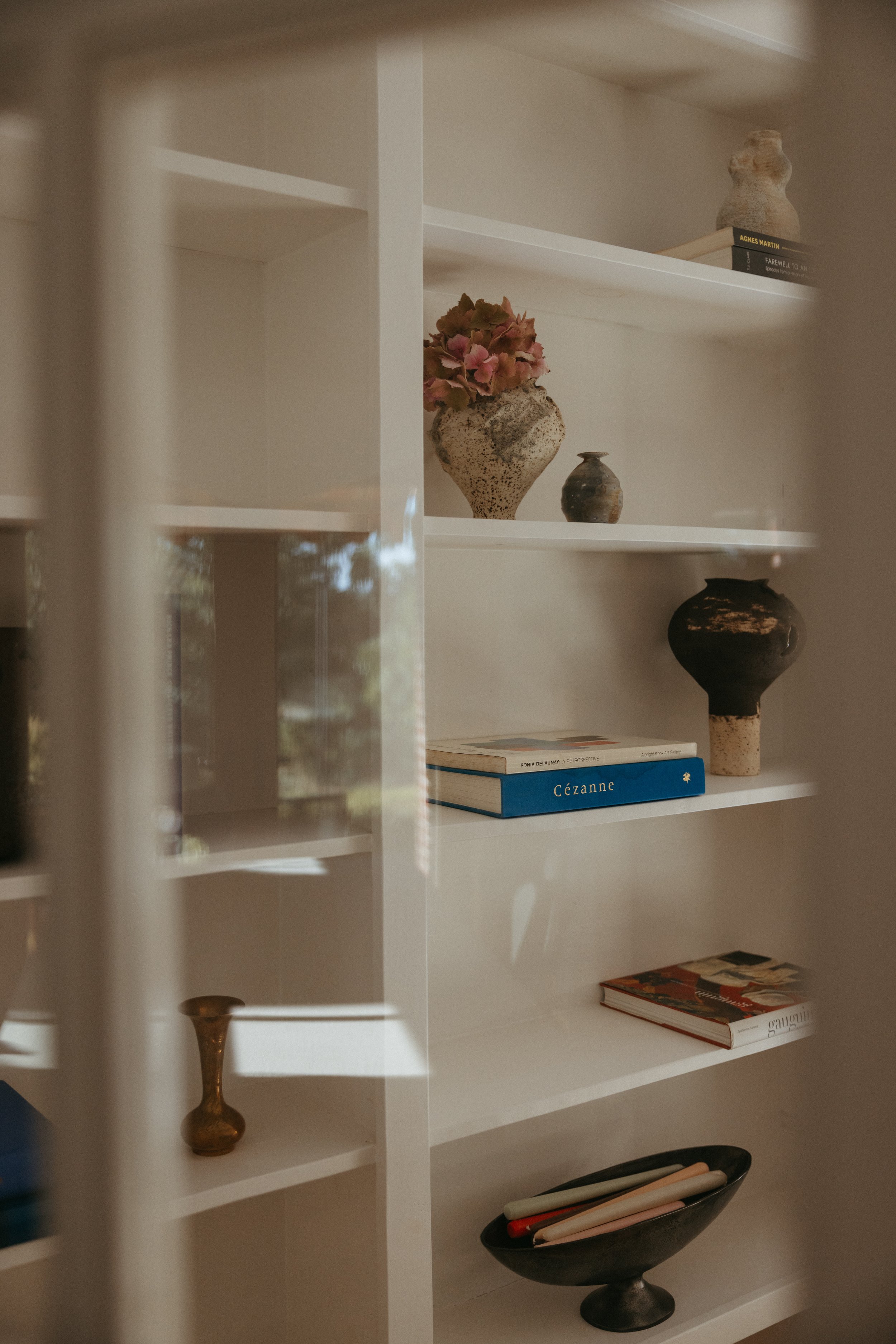 Unique ceramics and art books arranged on white shelves in a sunlit home interior with soft natural light.