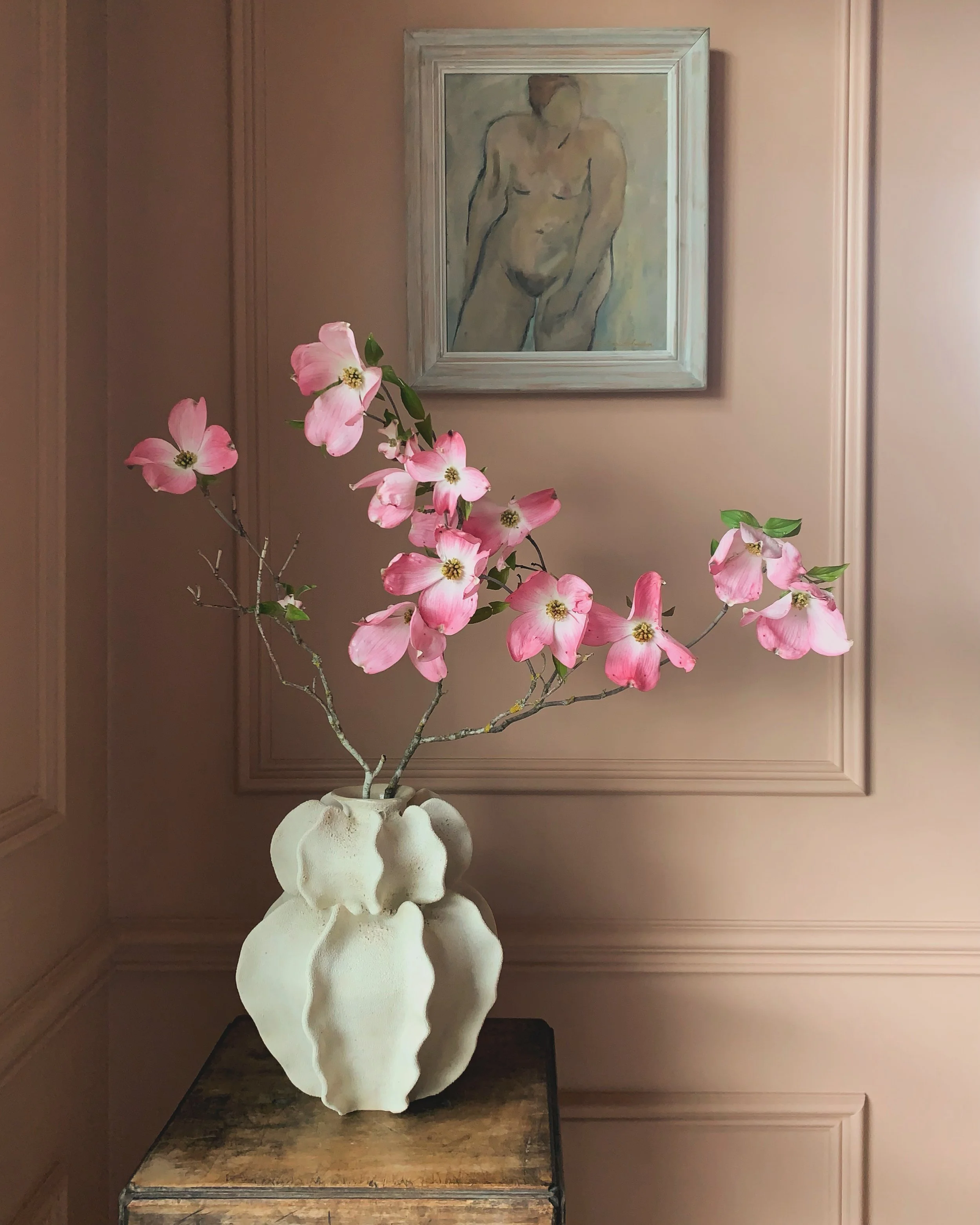 Soft pink dogwood branches arranged in a sculptural handbuilt ceramic vase, set against a blush-toned wall with a framed oil painting of an abstract nude figure—reflecting Ruby Bell Studio’s vintage fine art and custom ceramic aesthetic.