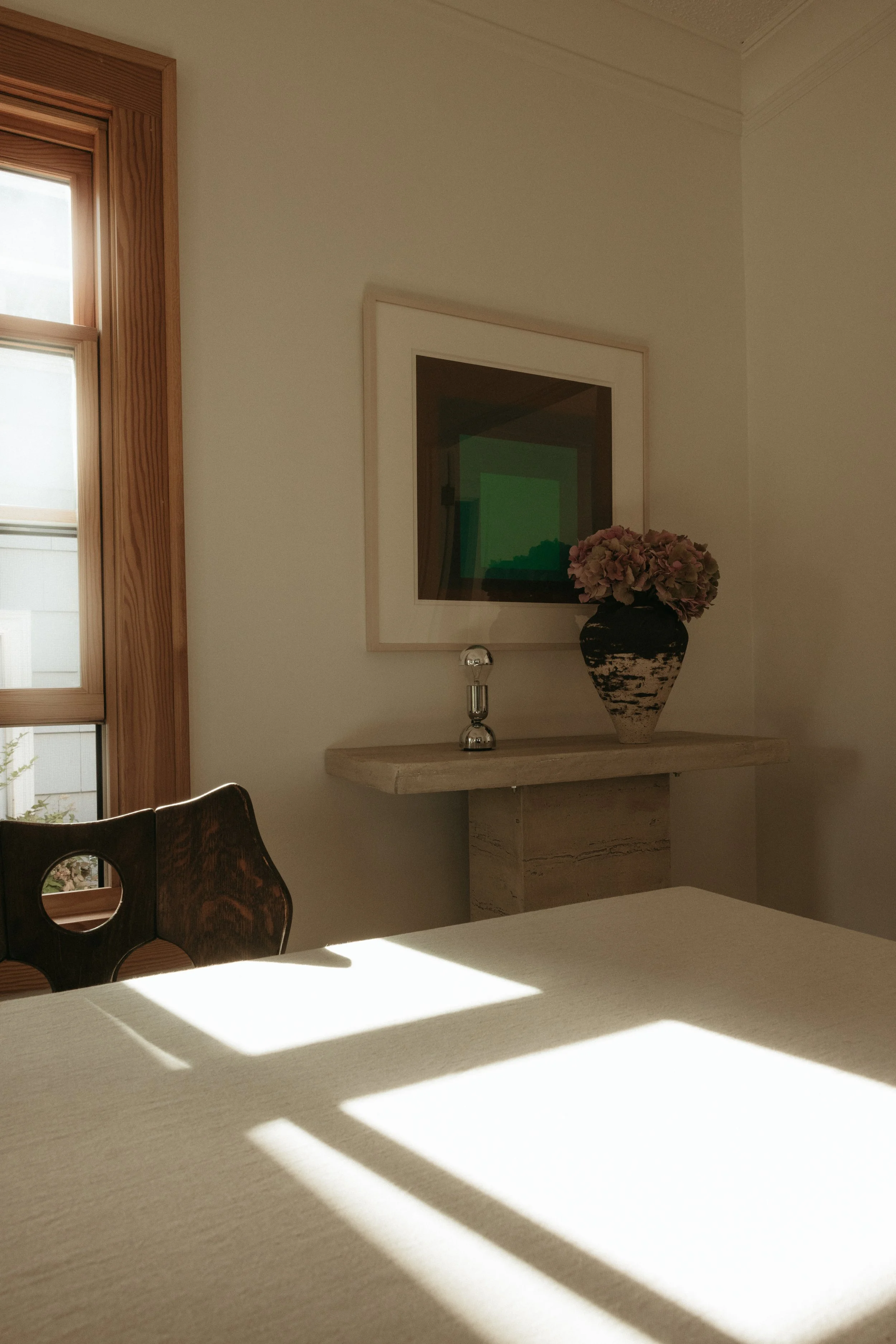 A dining room corner featuring a wooden window frame, a light-colored table with shadow patterns, a dark wooden chair, a marble console table with a black vase of pink hydrangeas, a modern metallic lamp, and a framed abstract picture on the wall.