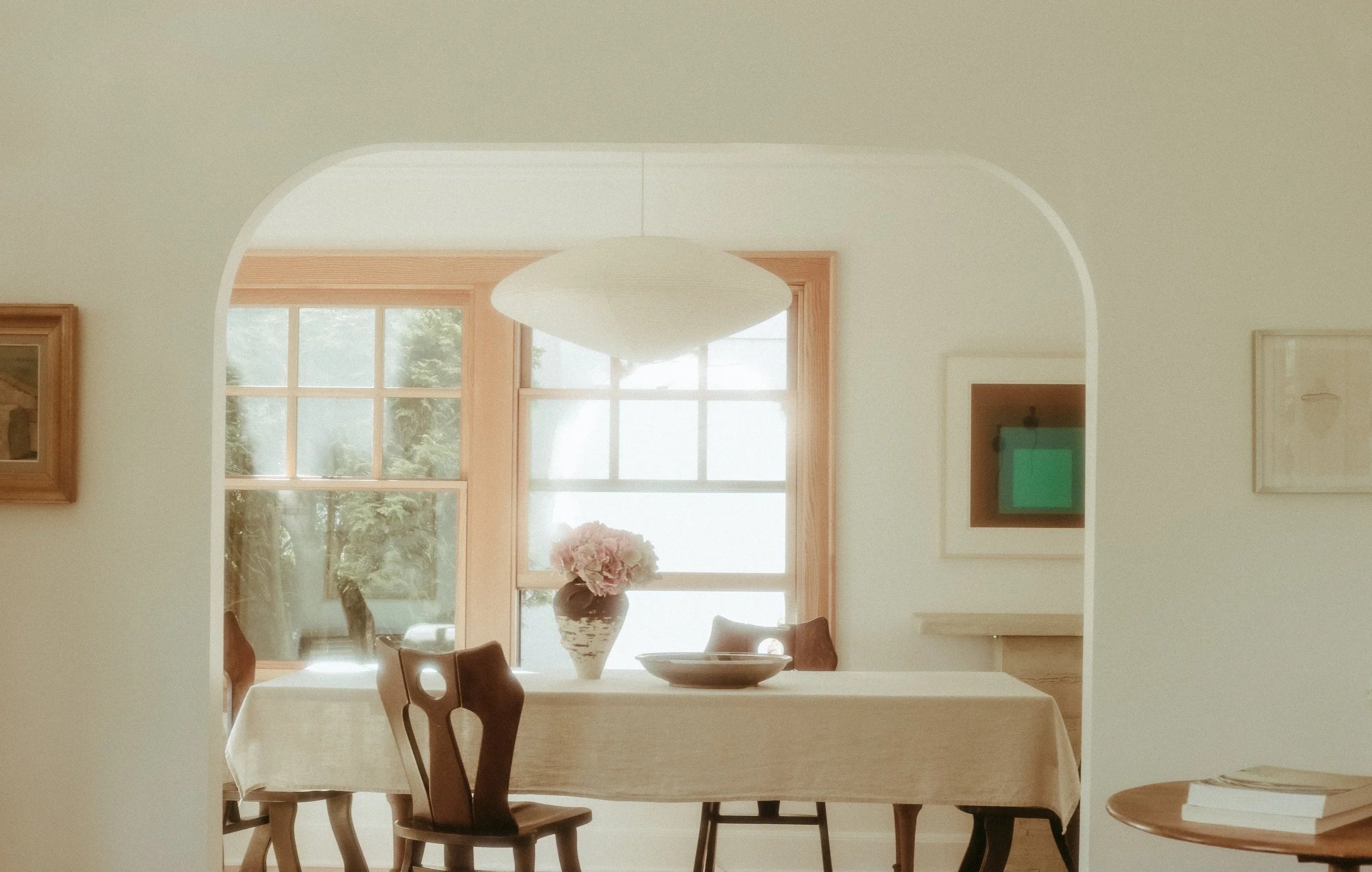 Soft, light-filled dining room featuring vintage wooden chairs, a linen-draped table, ceramic vase with flowers, and curated artwork
