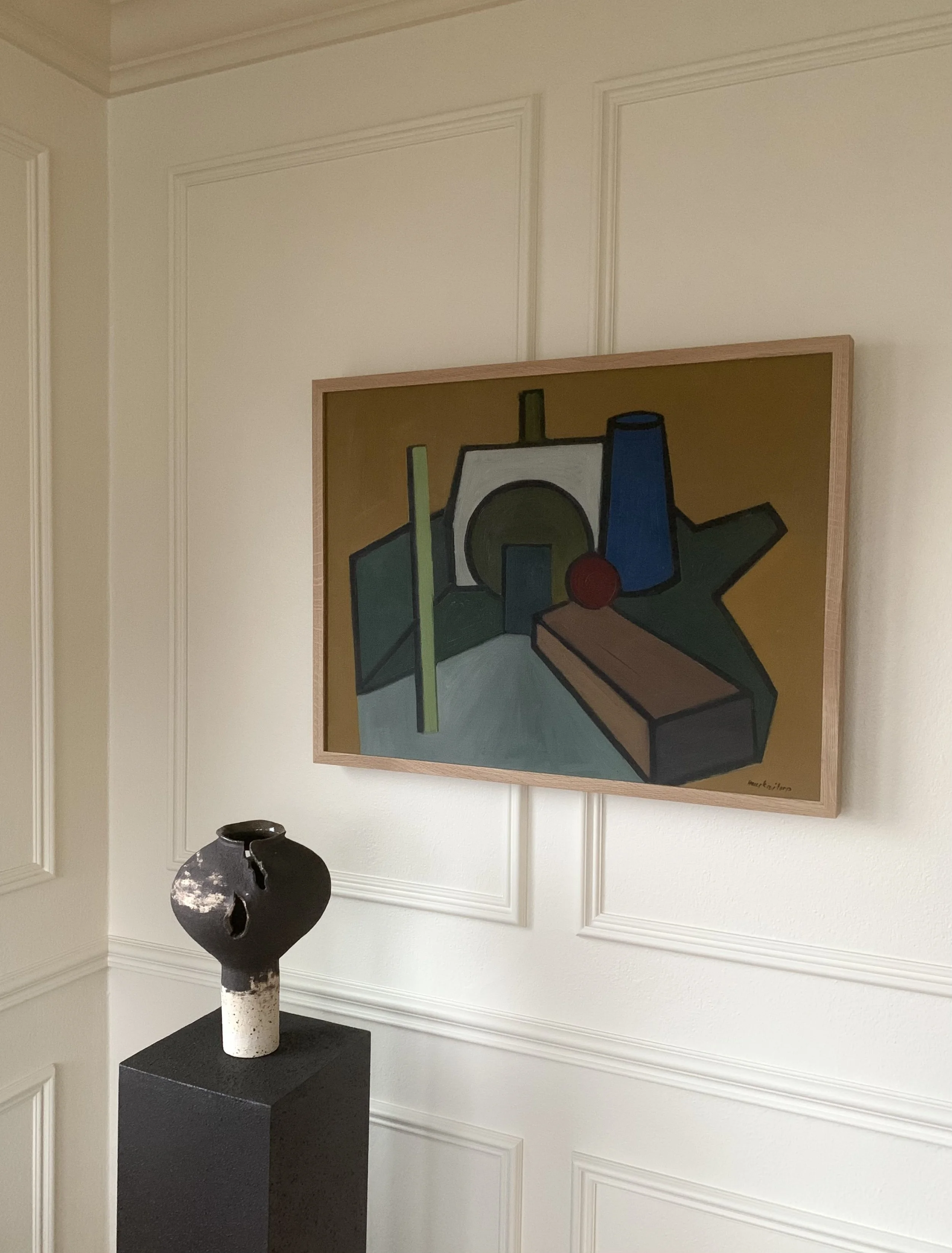 A marbled handbuilt Ruby Bell Studio vase on a pedestal in a paneled room, with a modern geometric still-life painting hanging above