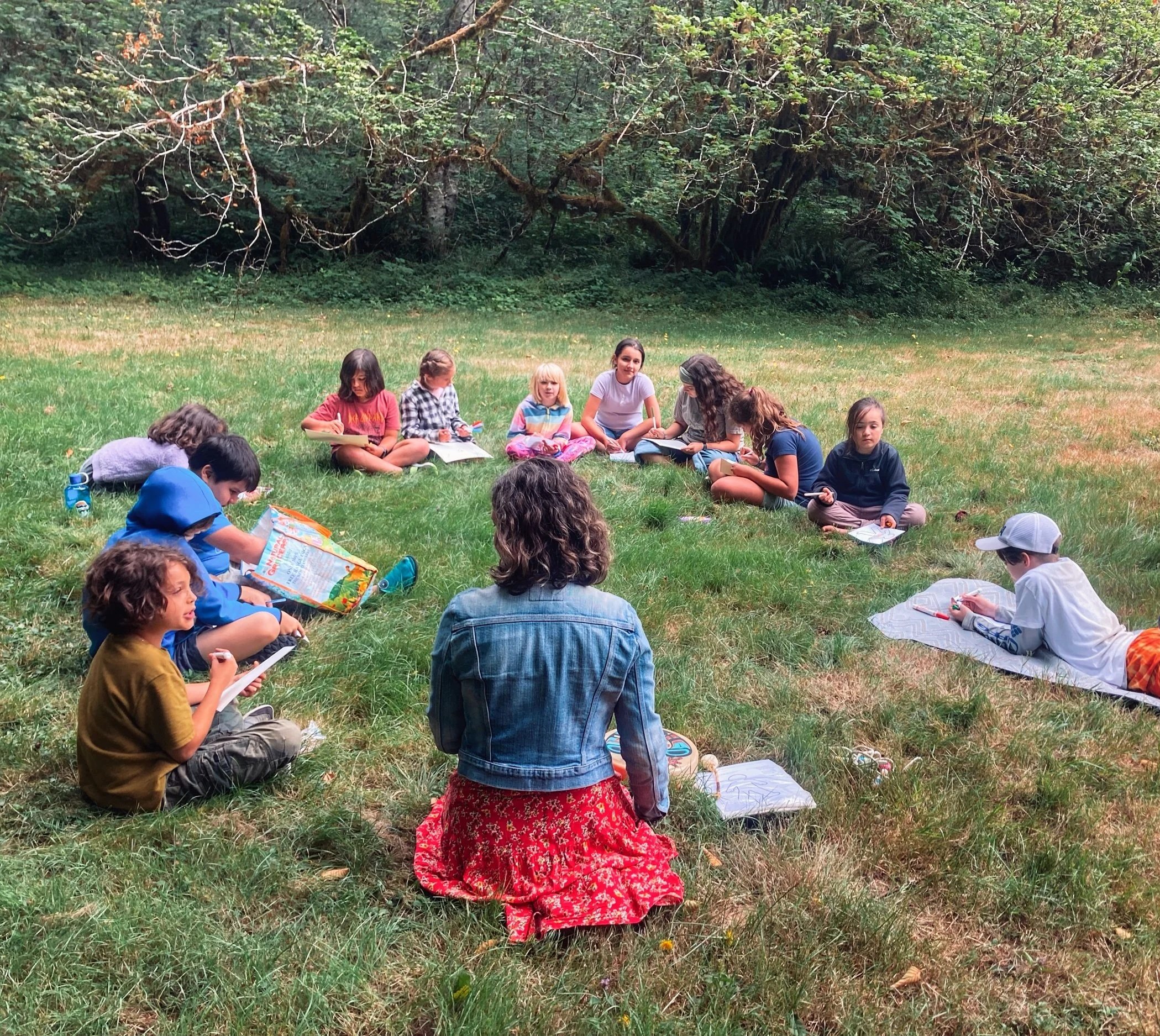 A group of children sitting on grass in a circle with Jenifer Trivelli outdoors, engaging in a mindful art activity near trees.