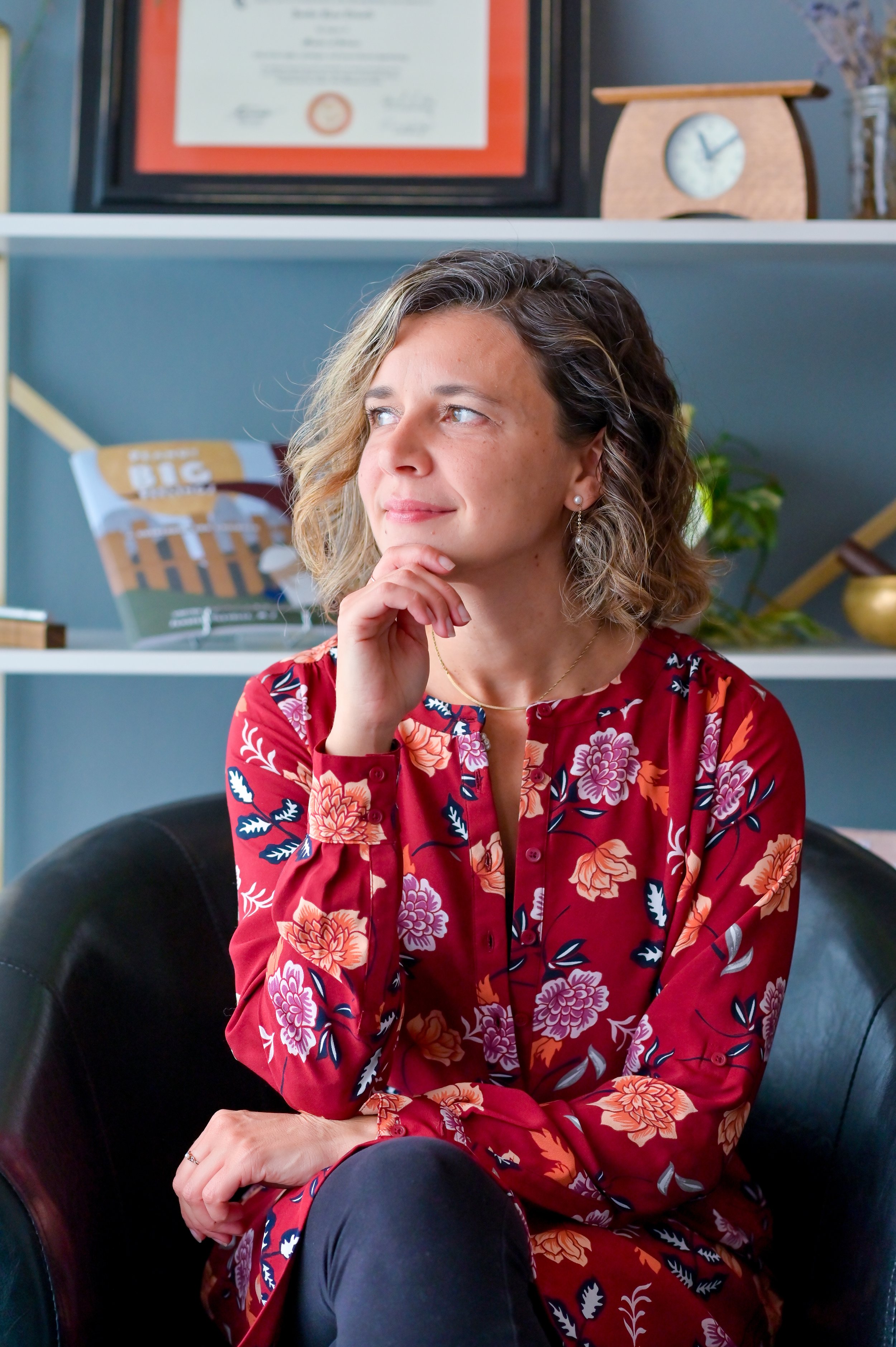 Jenifer Trivelli, a woman with curly hair and light skin, wearing a red floral dress, sits in a black chair with her chin resting on her hand, thoughtful. Behind her, a shelf displays a framed certificate, a clock, a plant, and decorative objects.