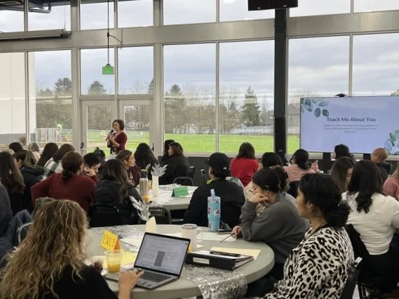 Jenifer Trivelli, a woman giving a presentation in a large, bright room filled with seated attendees, some taking notes or using laptops, with large windows showing an outdoor landscape.