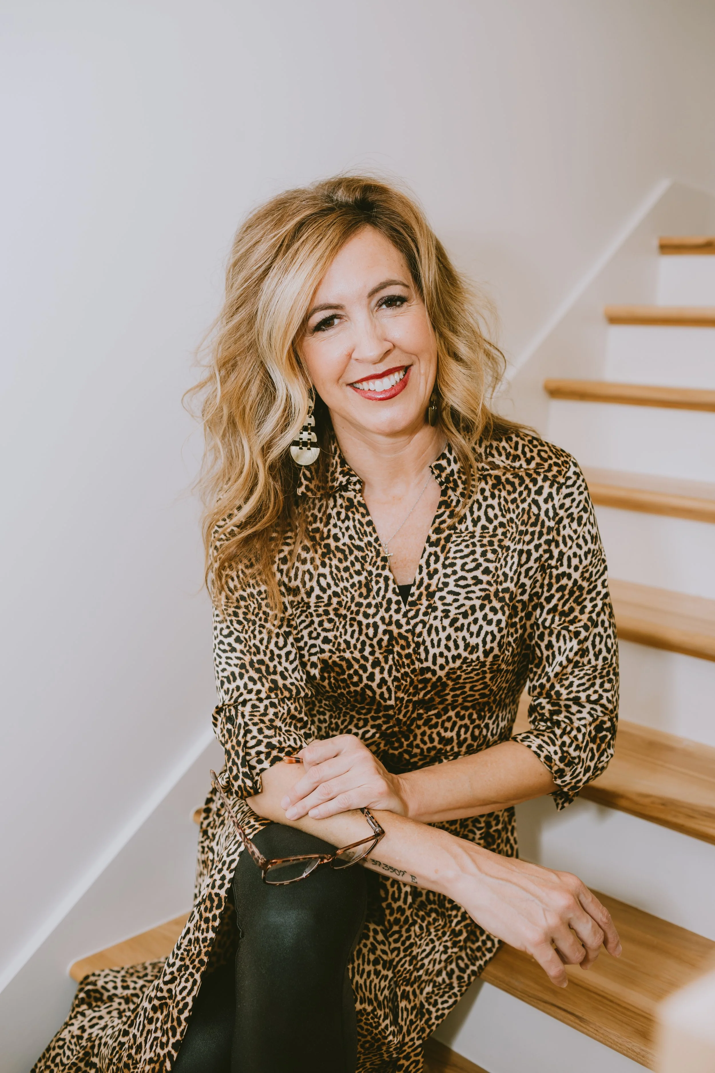 A smiling woman with wavy blonde hair, wearing a leopard print shirt and earrings, sitting on stairs and holding eyeglasses
