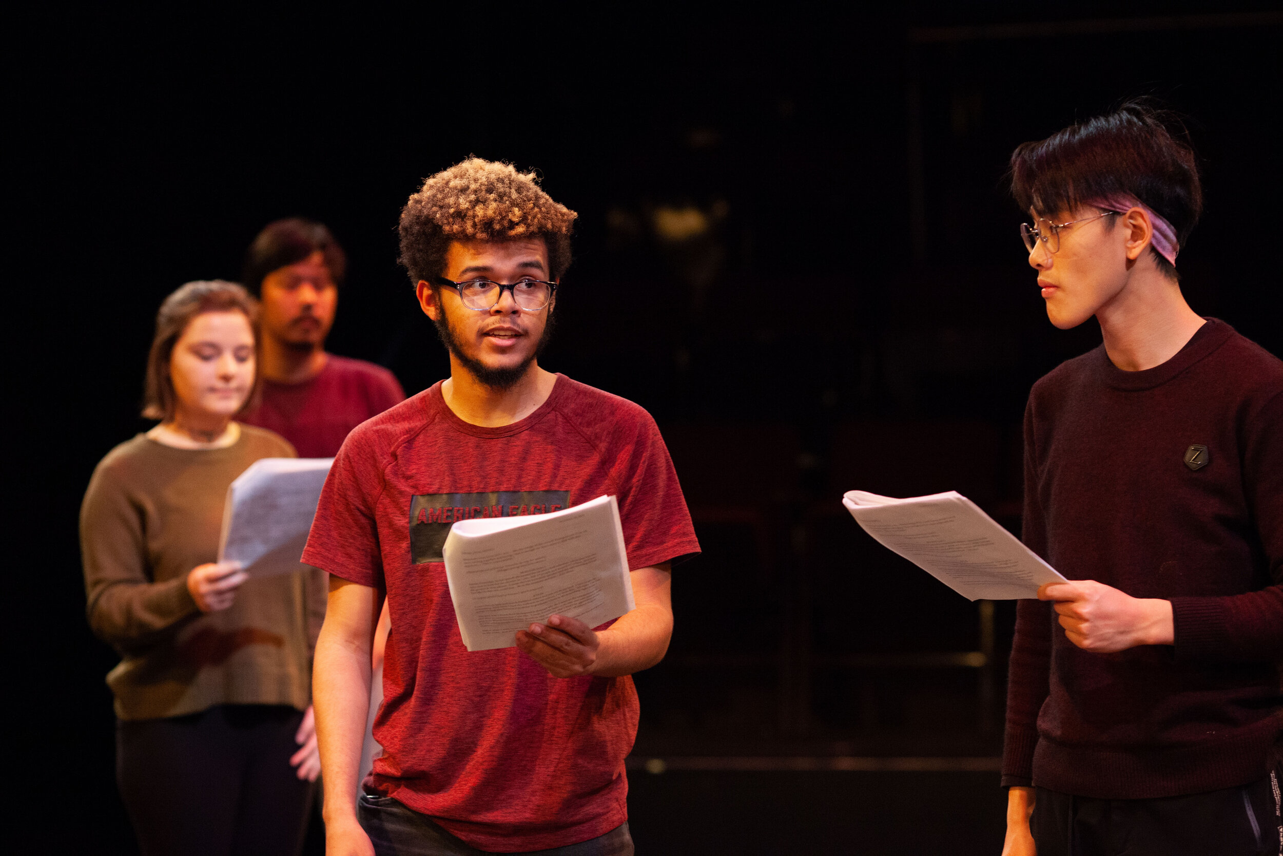  An actor with black and brown curly hair and a red shirt holds a script facing an actor with dark hair and a black shirt holding a script. 