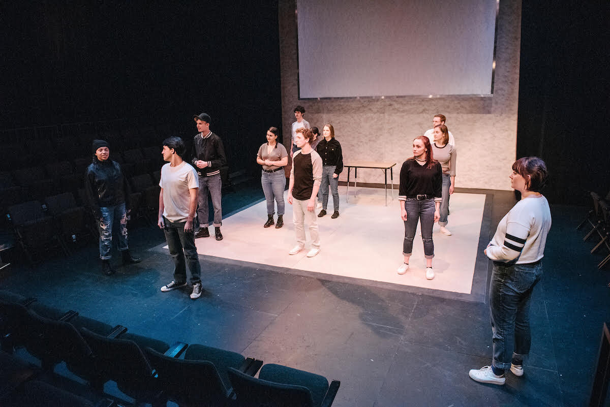  A cast of actors wearing gray and black shirts stand on a black stage with a white platform. 