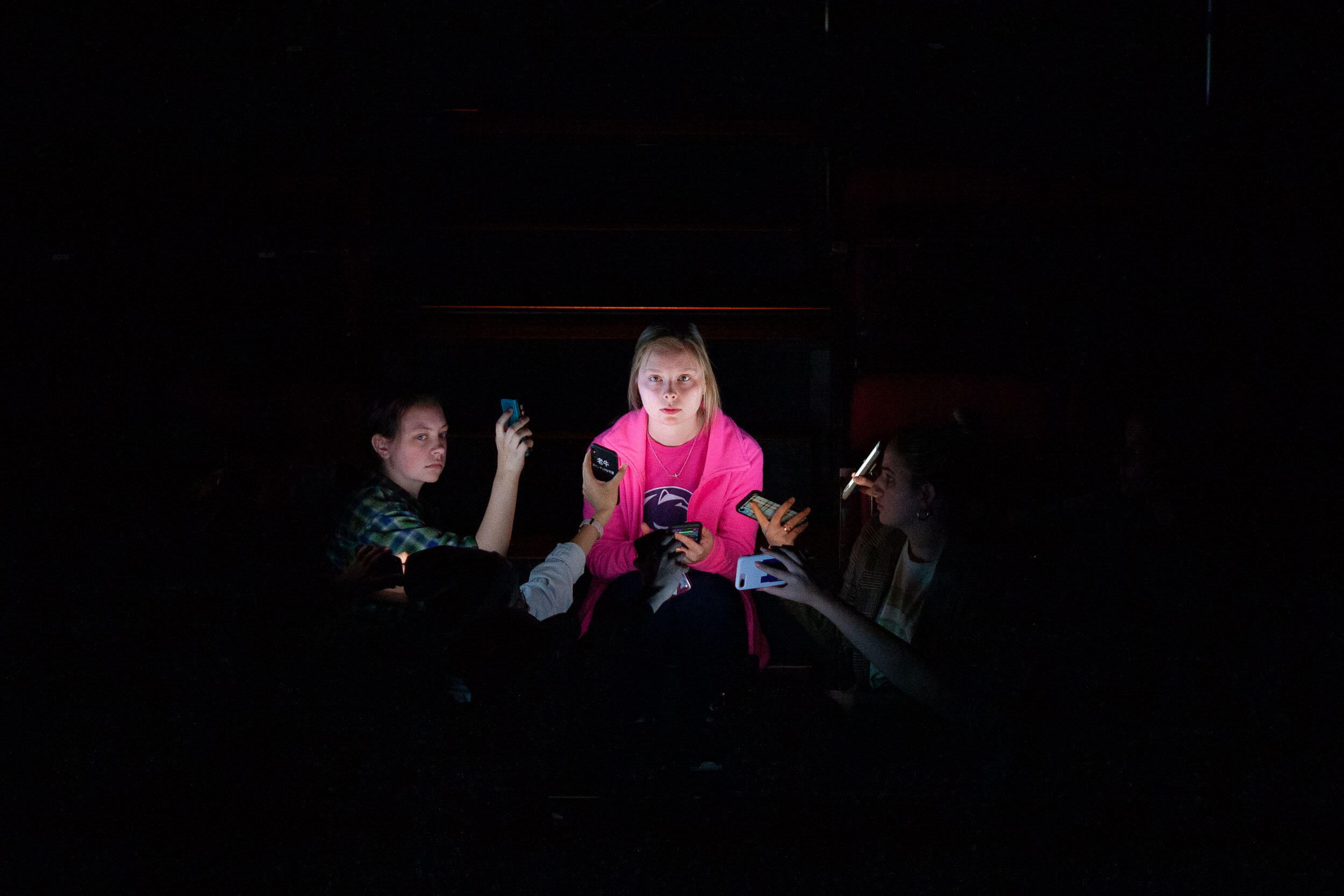  An actor in a pink shirt is illuminated by hands of other actors holding phones. 