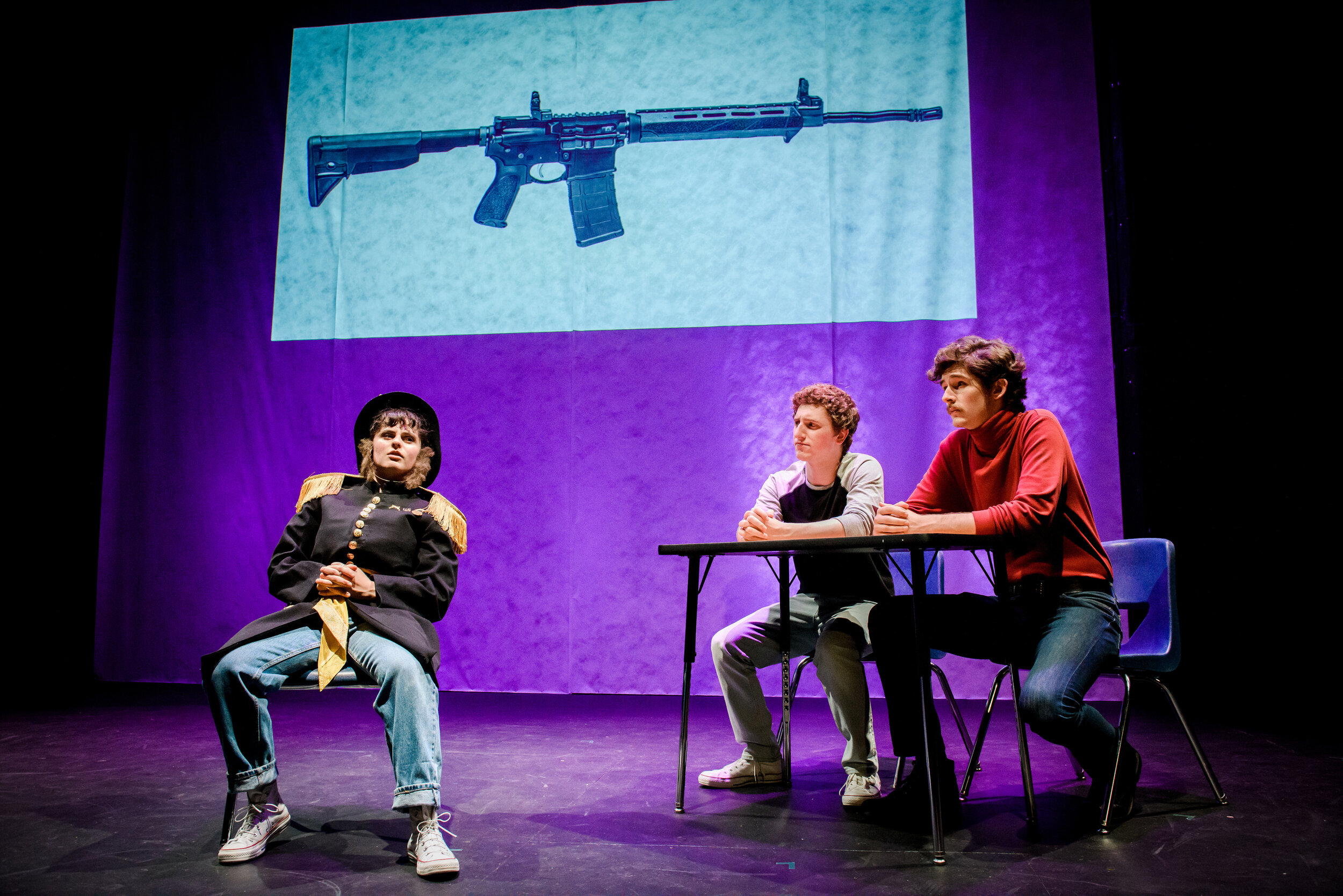  Three actors are on stage in front of a purple lighted projection with an image of an assault weapon on top. One actor is sitting, wearing an old-timey military jacket and the other two actors sit at a table. 