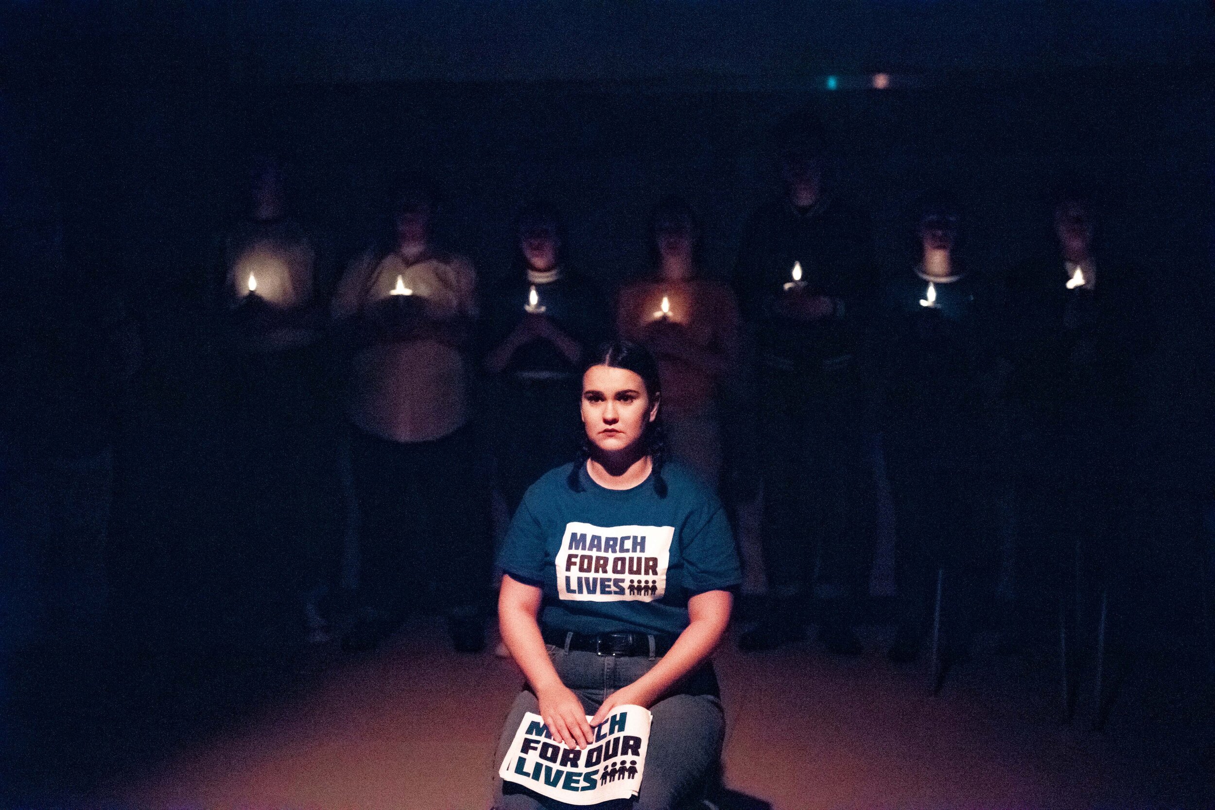  An actor wearing a blue shirt with the March for Our Lives logo and holding a March for Our Lives poster sits on their knees in front of a line of shadowy actors holding small candles. 