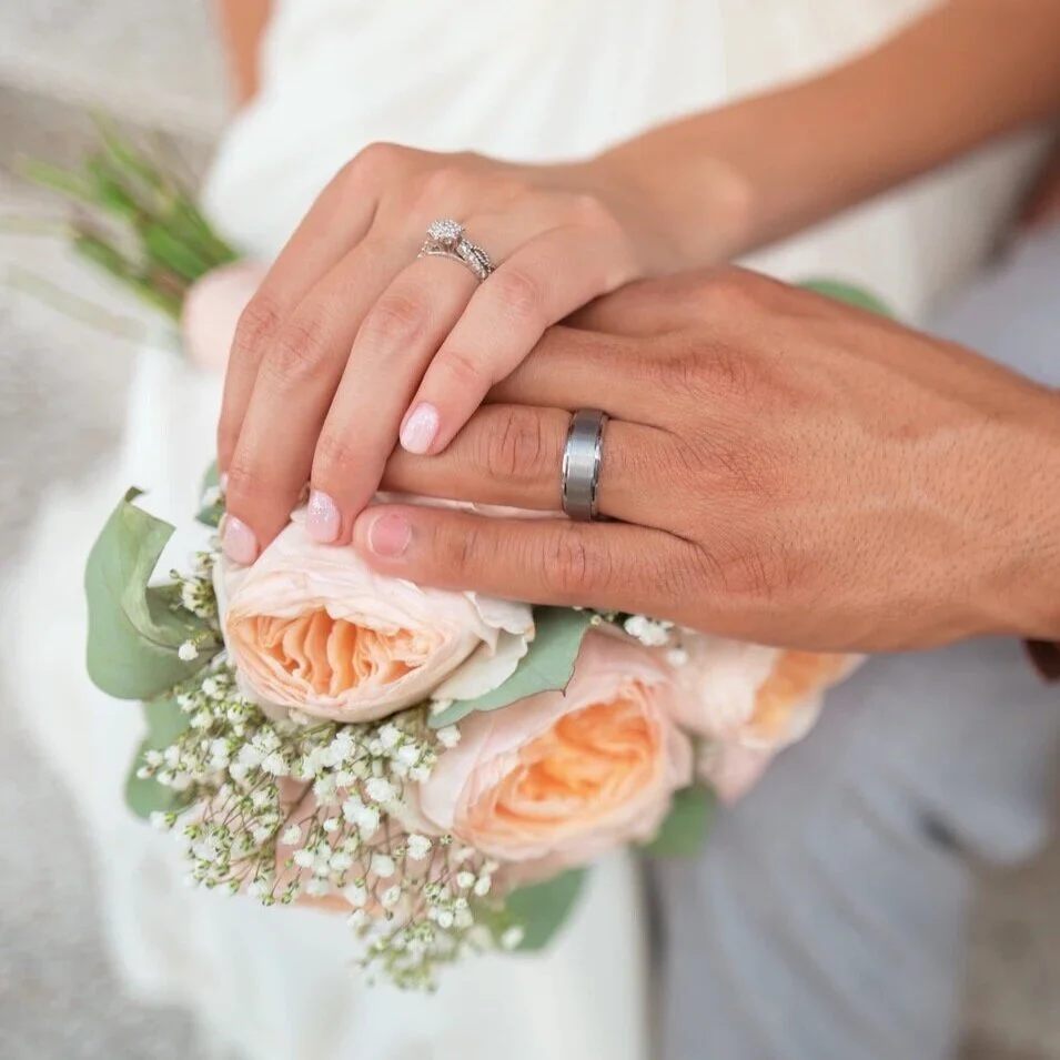 Close-up of a newlywed couple's hands wearing wedding rings, resting on a bouquet of light pink and white roses with greenery.