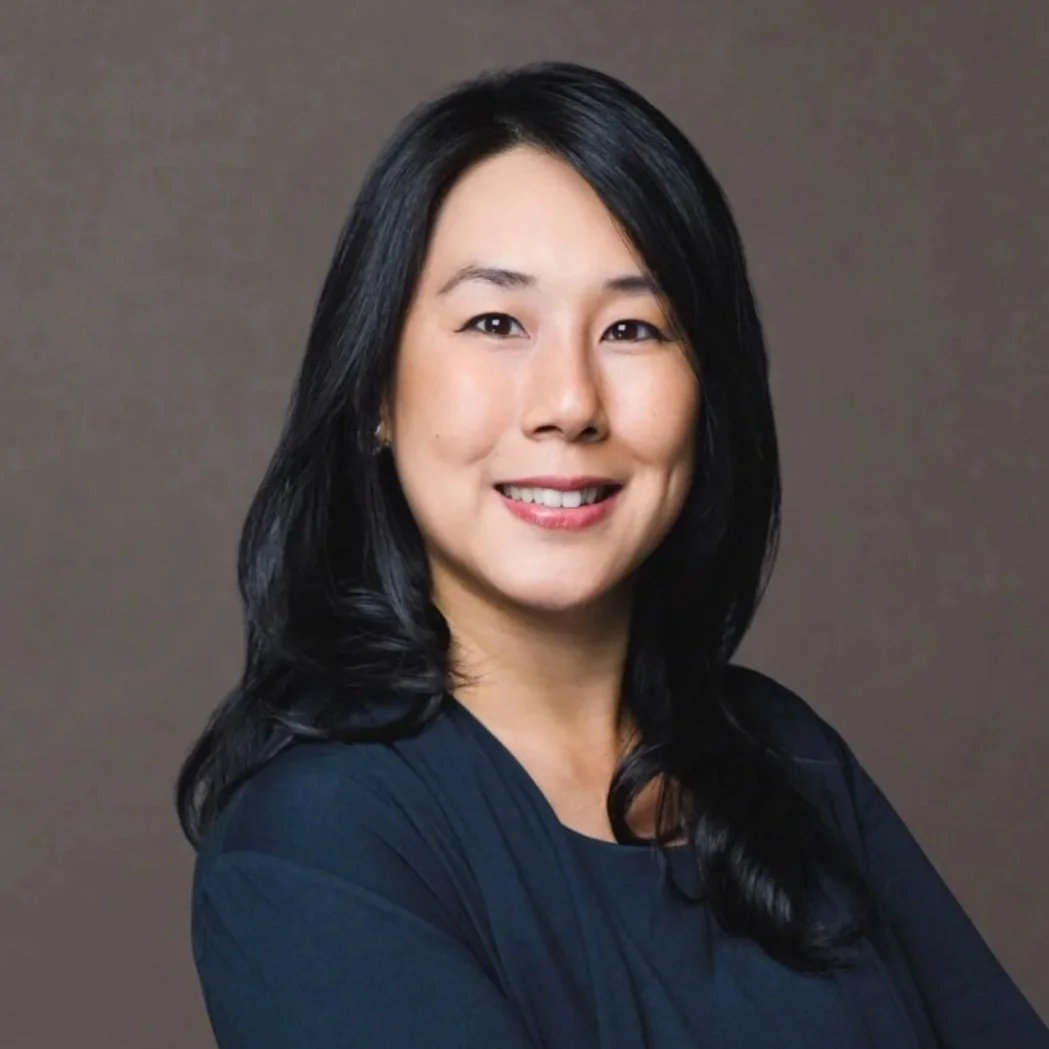 A woman with long black hair, smiling, wearing a black top, in front of a gray background.