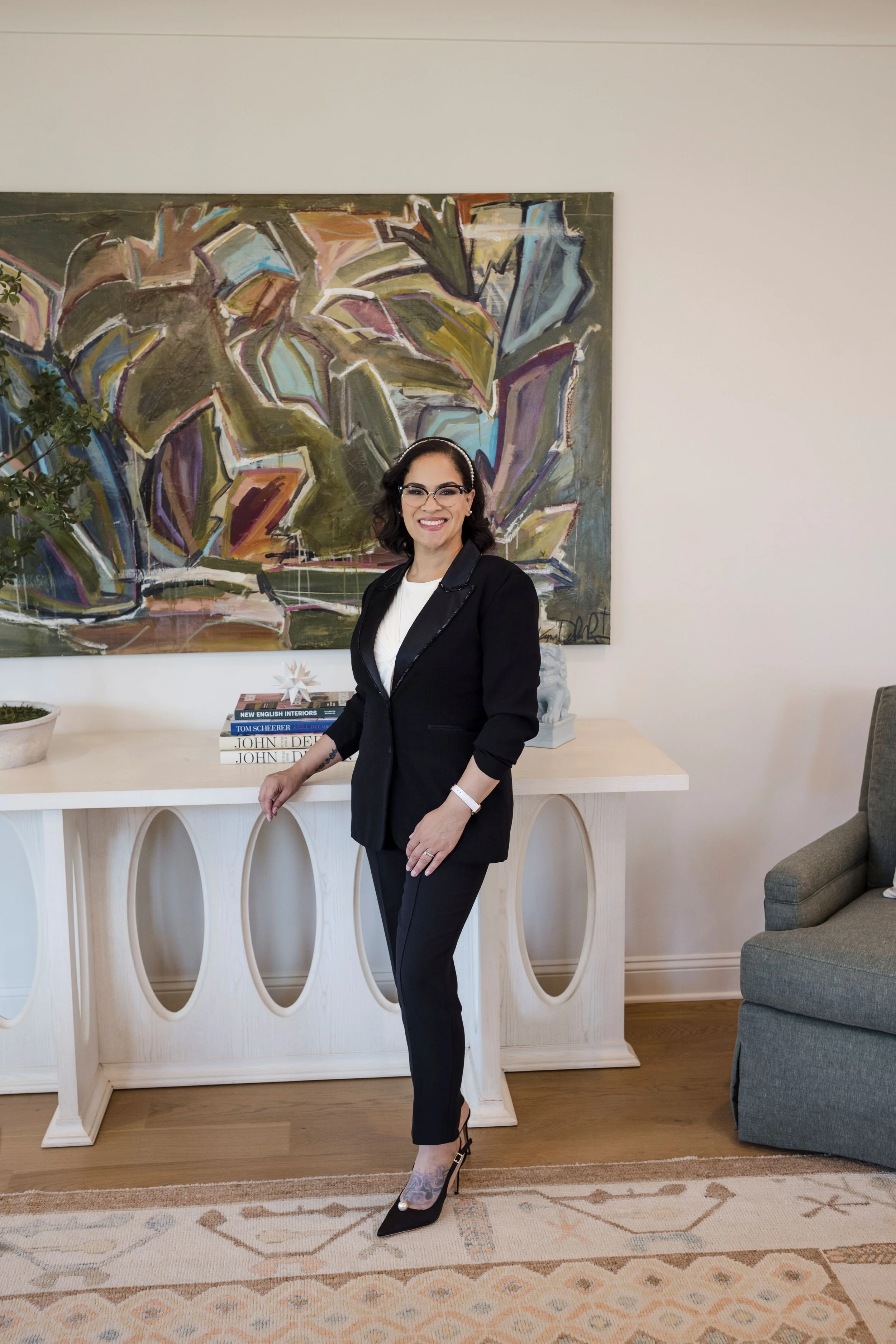 Woman in black suit with glasses smiling in a modern living room with an abstract painting behind her on the wall, a stack of books on a white decorative table, and a gray armchair to the right.