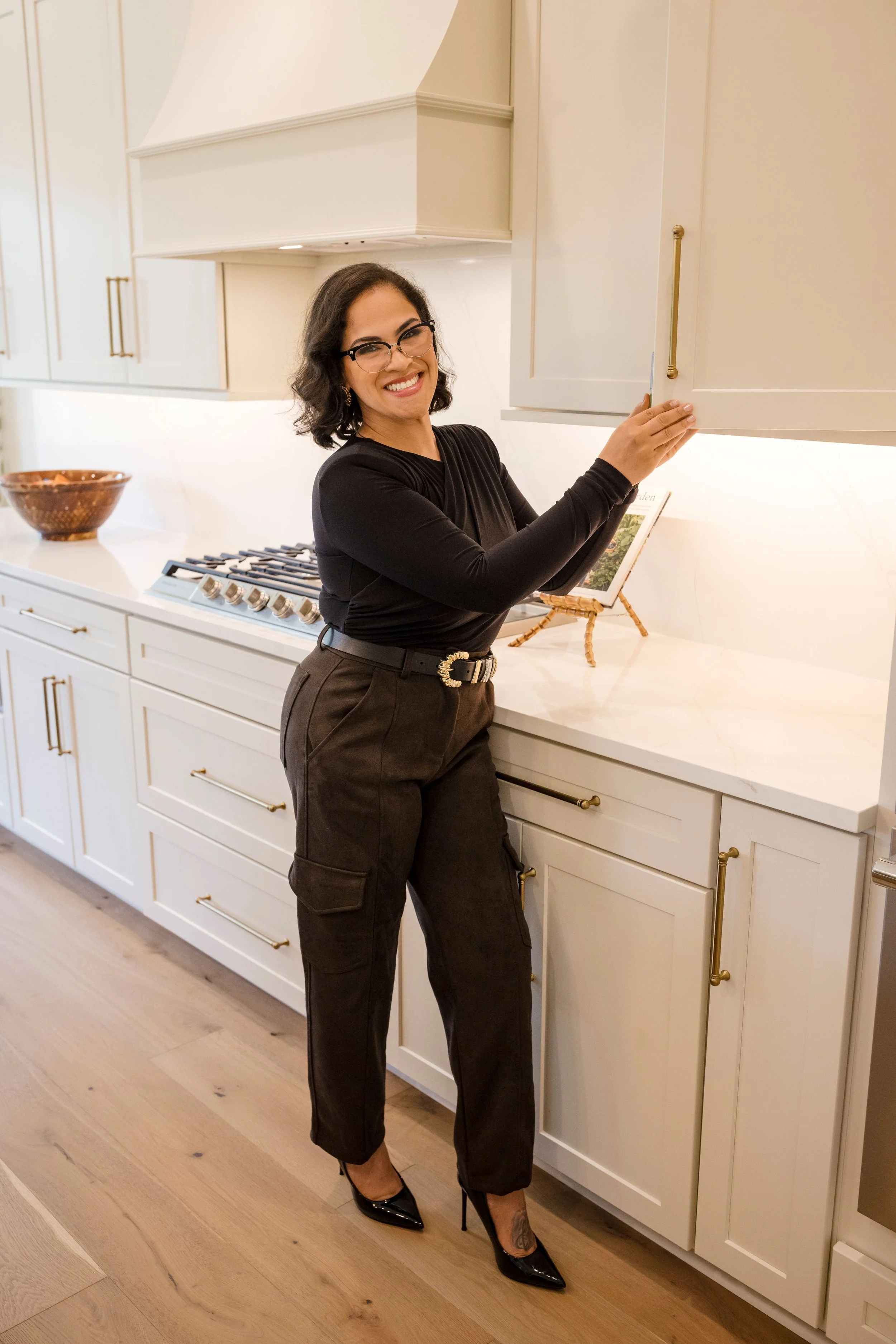 Woman with dark hair and glasses smiling in a modern kitchen with white cabinets and gold handles, wearing a black long sleeve top and dark pants, standing with one hand on the cabinet.